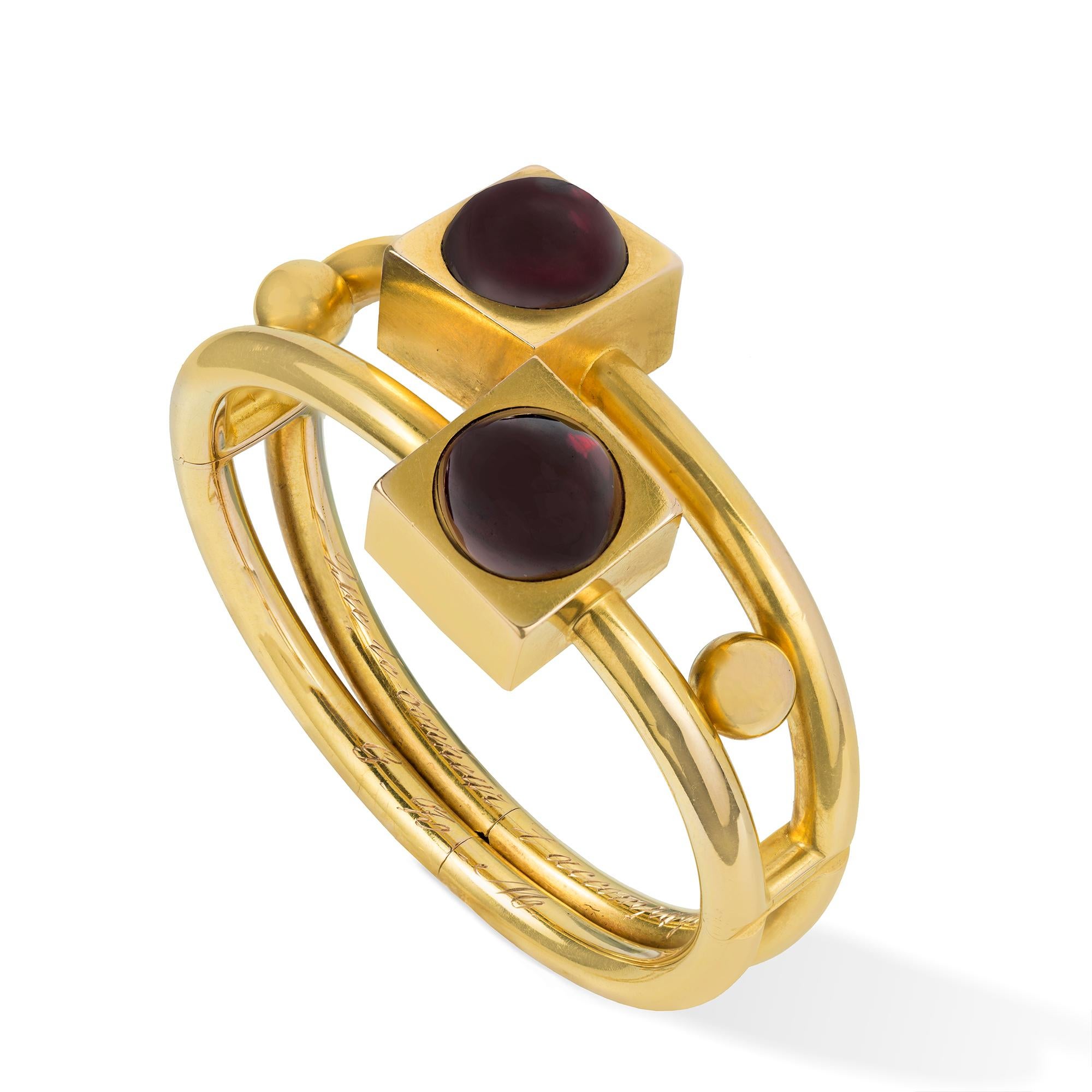 A late nineteenth-century gold and garnet bangle, the geometrical designed bangle centrally with the two gold cubes each set with a round carbuncle garnet, to a split yellow gold hinged bangle with concealed hinged clasp and safety chain, all in