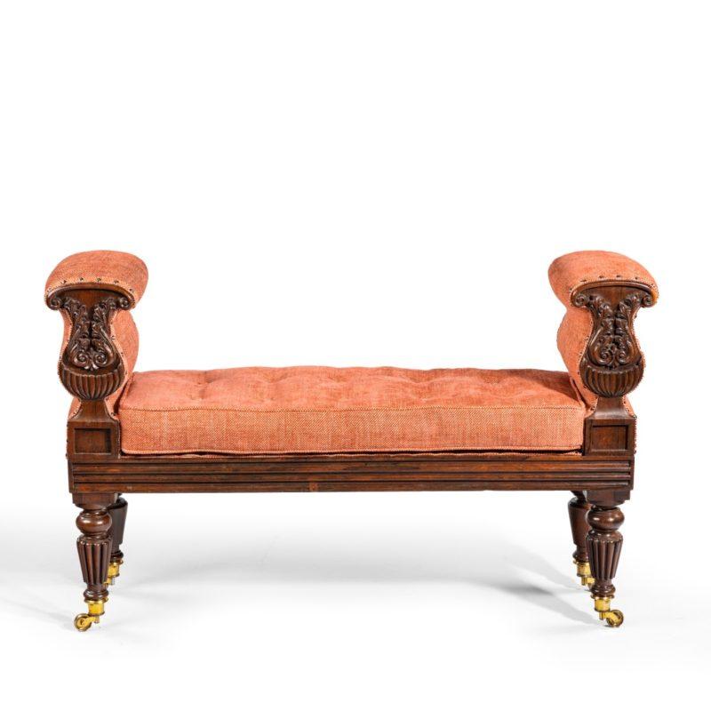 A late Regency rosewood window seat, in the manner of George Oakley, of rectangular form, with vase-shaped, upholstered arms, reeded seat rails and tapering gadrooned legs with the original brass castors, the arms carved with bold acanthus scrolls,