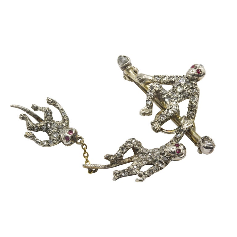A late Victorian fun monkey bar brooch, the bar set with rose-cut diamonds either end with a monkey sitting in the centre with rose-cut diamond set body and ruby set eyes, with one hand holding a similarly decorated monkey hanging from the bar, with