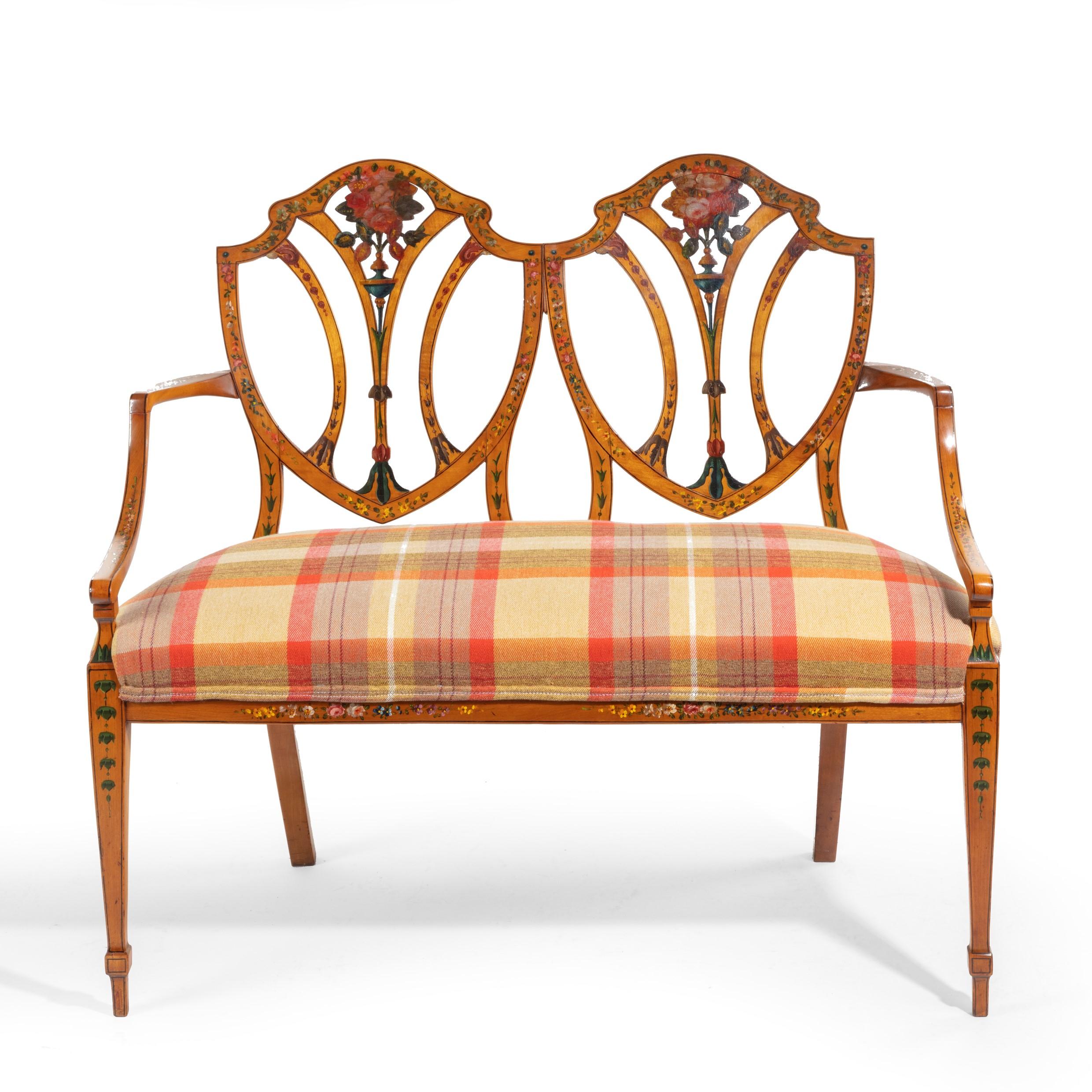 A late Victorian Sheraton revival painted satinwood two-seat settee, the double shield back and curved arms set upon square tapering front legs and outswept back legs, painted in polychrome with a central roses and floral garlands, reupholstered.