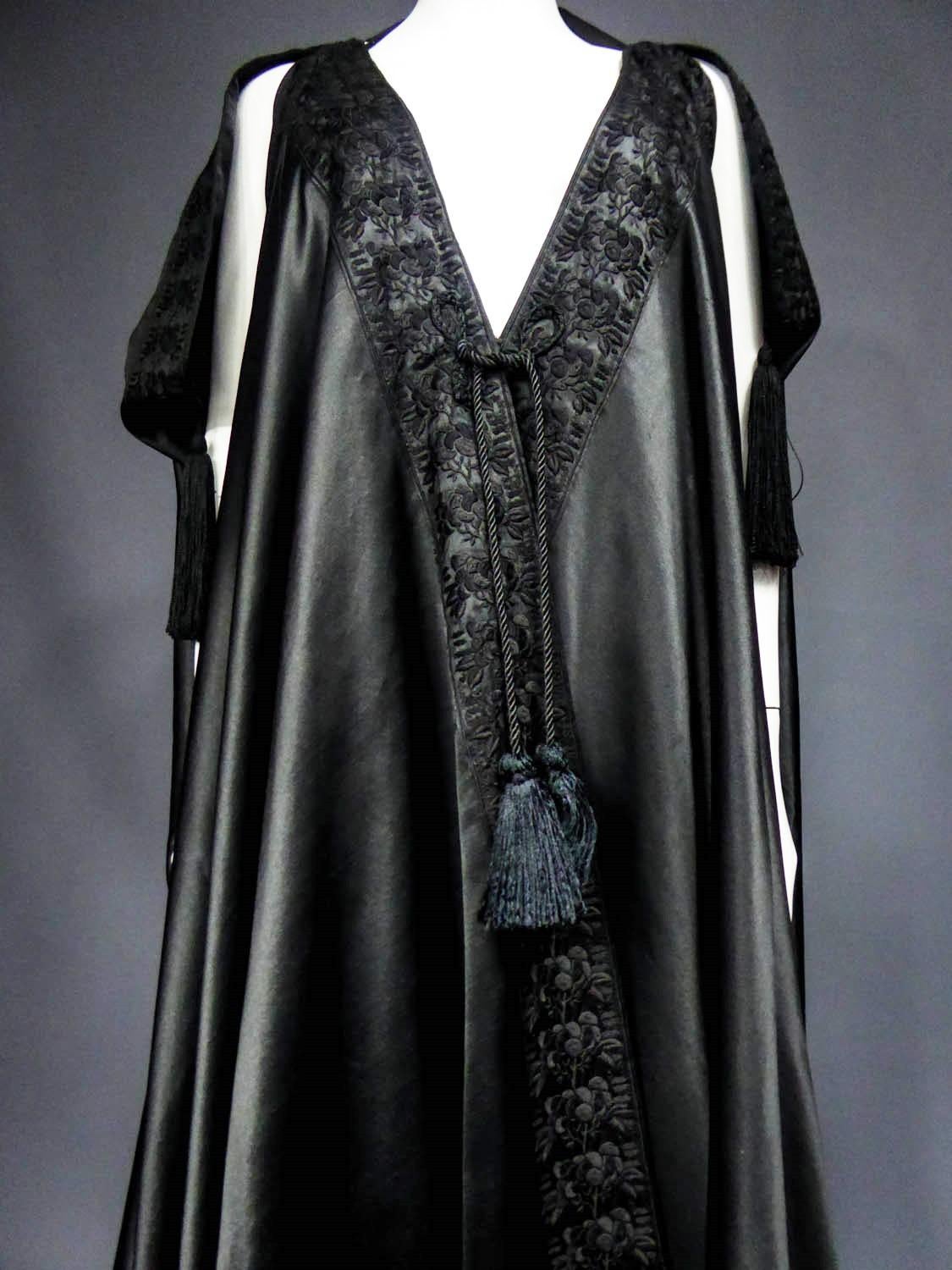Circa 1920
England

Amazing evening cape in the Djellaba spirit in black Duchesse satin silk by Liberty and Co dating from the years 1920 - 1930. Cape with small slashed sleeves and pompoms of silk trimmings to pass the arms. Beautiful black duchess