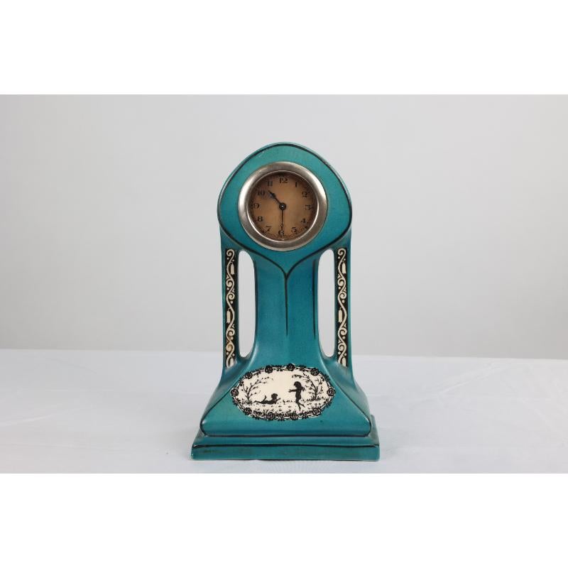 A little Art Nouveau turquoise blue dressing table clock decorated with little cherubs playing and lazing in a country scene within a ring of roses, and also bluebells hanging to the sides. 
