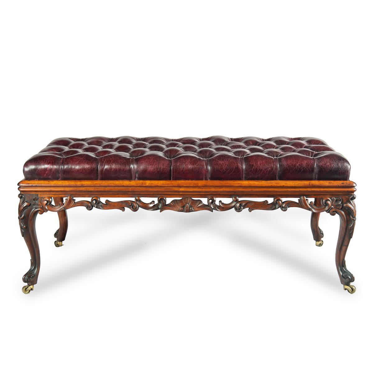 Taburete largo de palisandro de principios de la época victoriana, de forma rectangular sobre cuatro patas cabriolé con ruedas de latón,  el friso tallado con volutas foliadas caladas, el asiento abatible retapizado en cuero burdeos abotonado. 