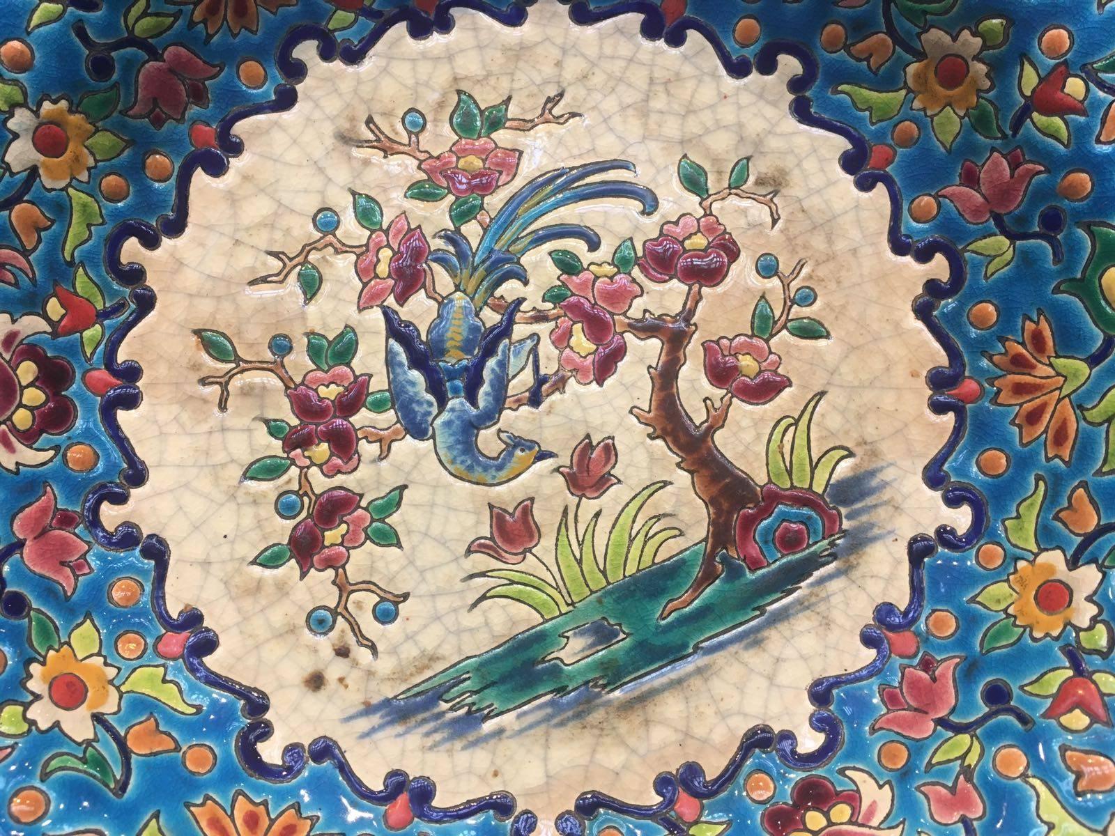 Made of French faience, the handmade shallow bowl features slightly scalloped edges and a foot. The top (inside) portion is enameled with a central 'medallion' scene of a bird perched atop a tree, that is surrounded by a border of a colorful