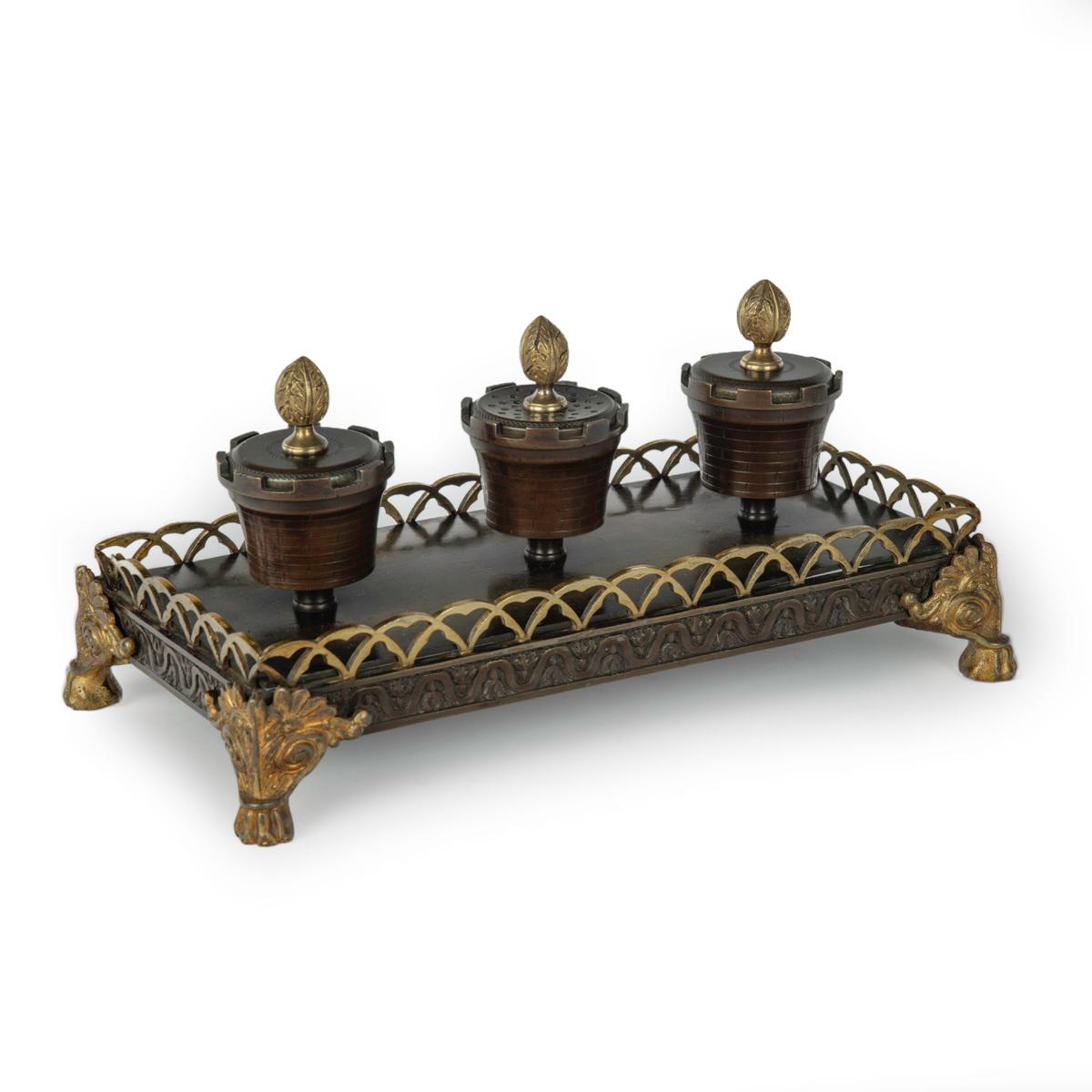 A Louis Philippe bronze and ormolu desk set, comprising two ink wells and a powder shaker in the form of crenelated urns with anthemion-bud finials, all set on a rectangular base with a  gallery and applied anthemion scroll and paw feet.  French,