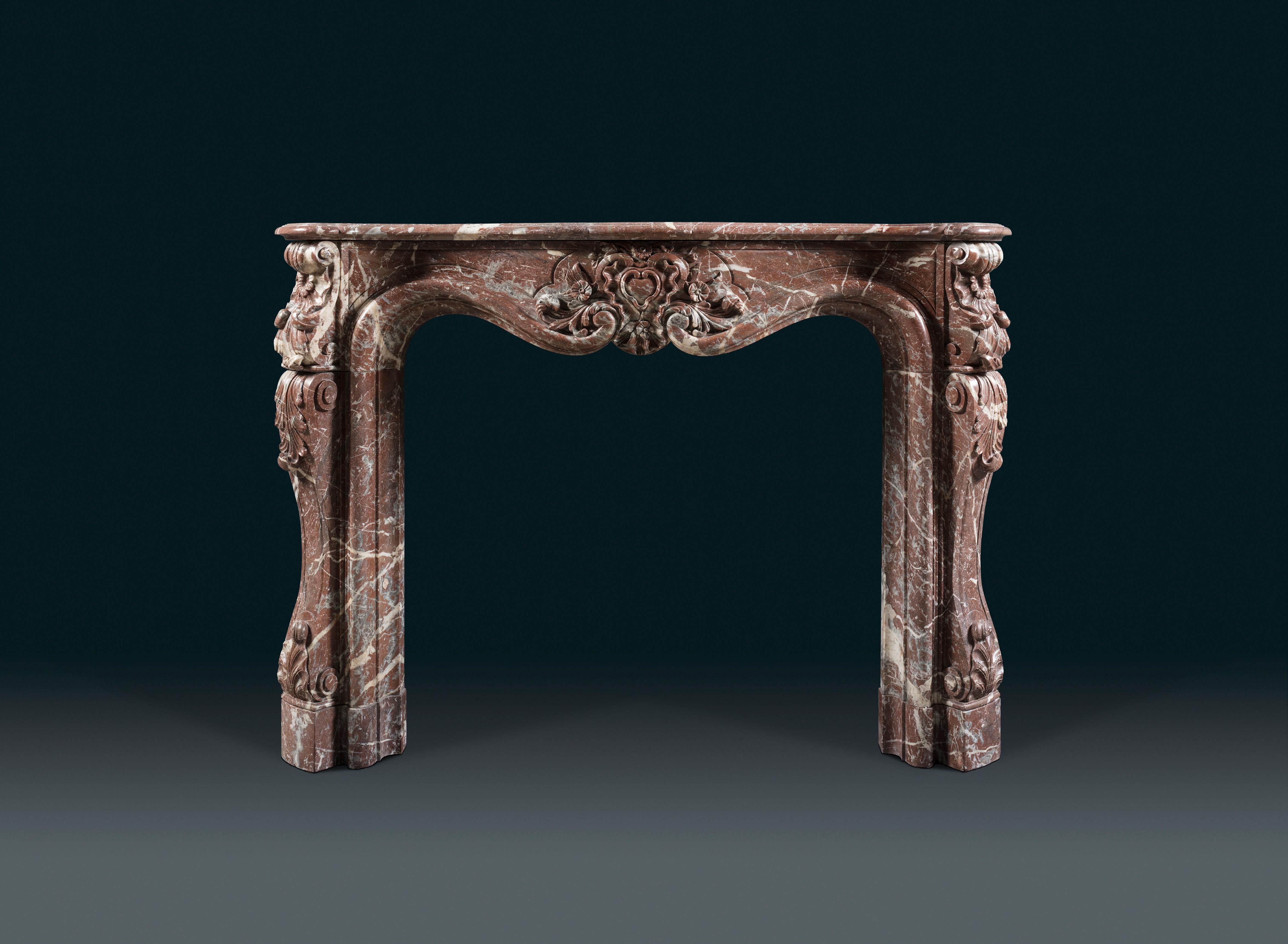 A Louis XV style Rococo chimneypiece in Rosso Antico marble with an elegant, moulded shelf. The fine asymmetric serpentine frieze presents a central cartouche in the shape of a stylised shell, flanked by foliage and panels. Shell corner blocks