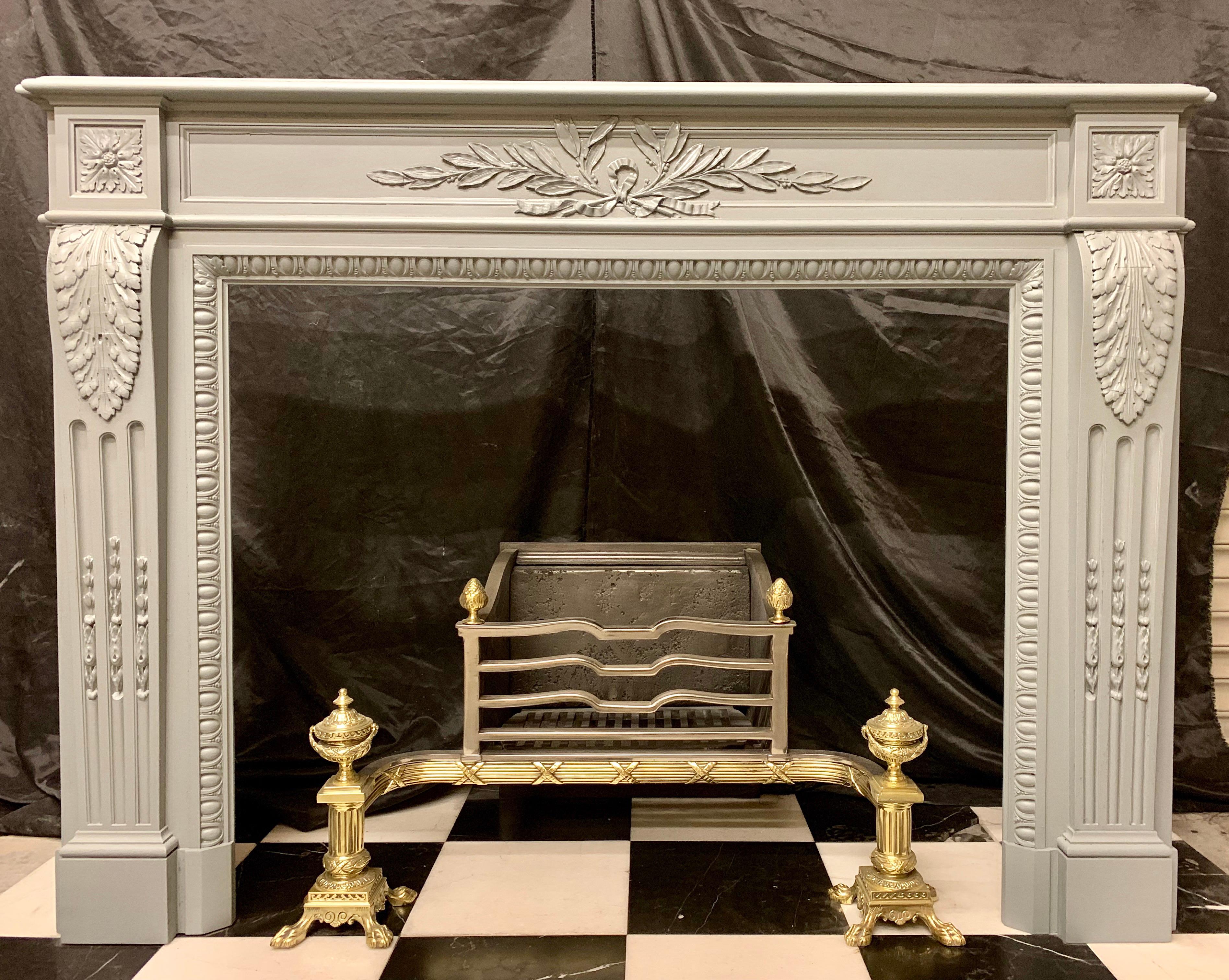 A Louis XVI style 19th century mahogany fireplace surround painted in French Grey. The breakfront shelf rests above a lambs tongue beaded frieze centred by a delicately carved ribboned, leaf, and berry wreath and sprigs, flanked on the end blocks by