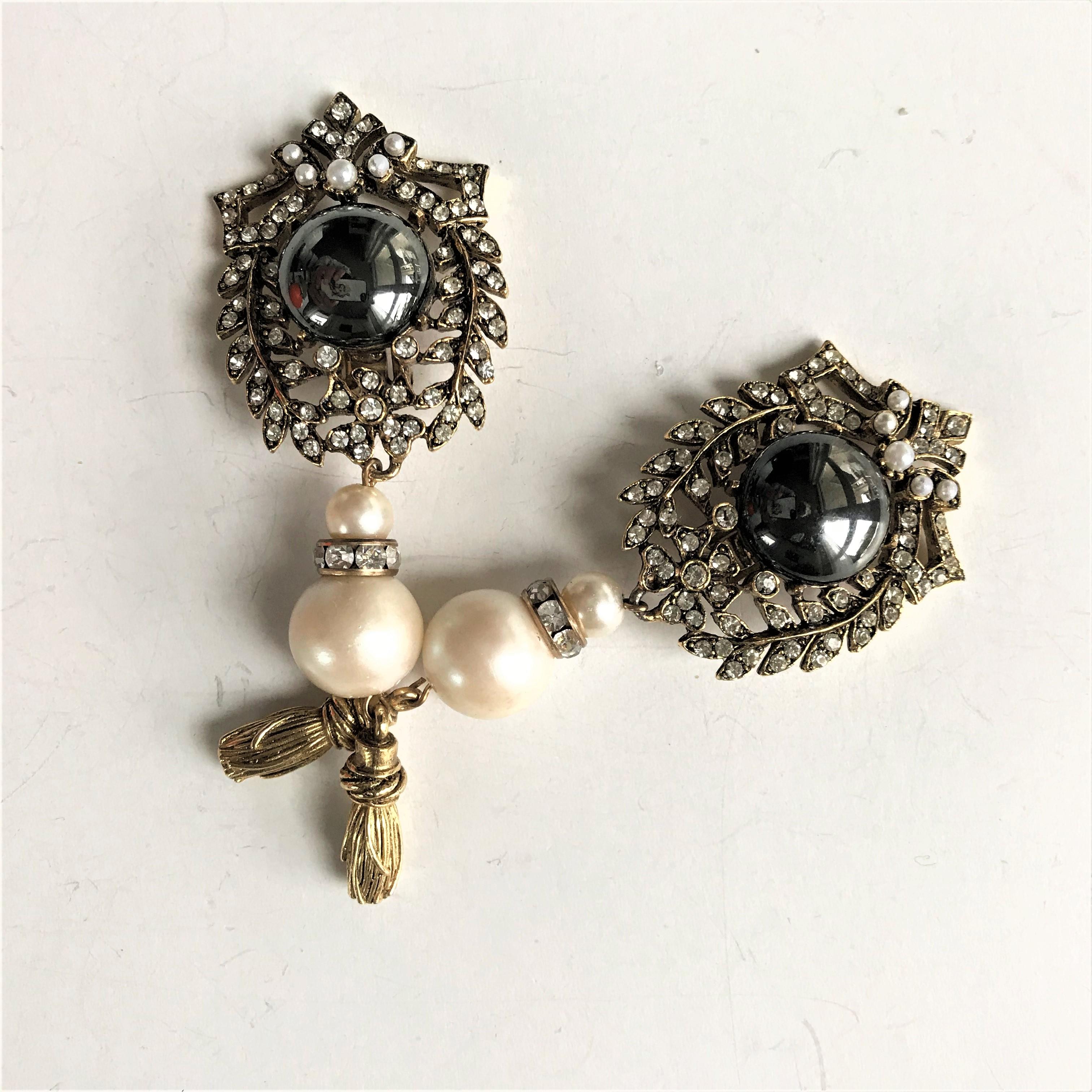 Pair of ear clip unsigned with a central shiny anthrazit stone, trimmed with fine leaf branches. Small rhinestones in the border. A false large pearl and a tassel are attached to it.

Dimensions length 10 cm, upper part 5 cm x 4 cm, attached large