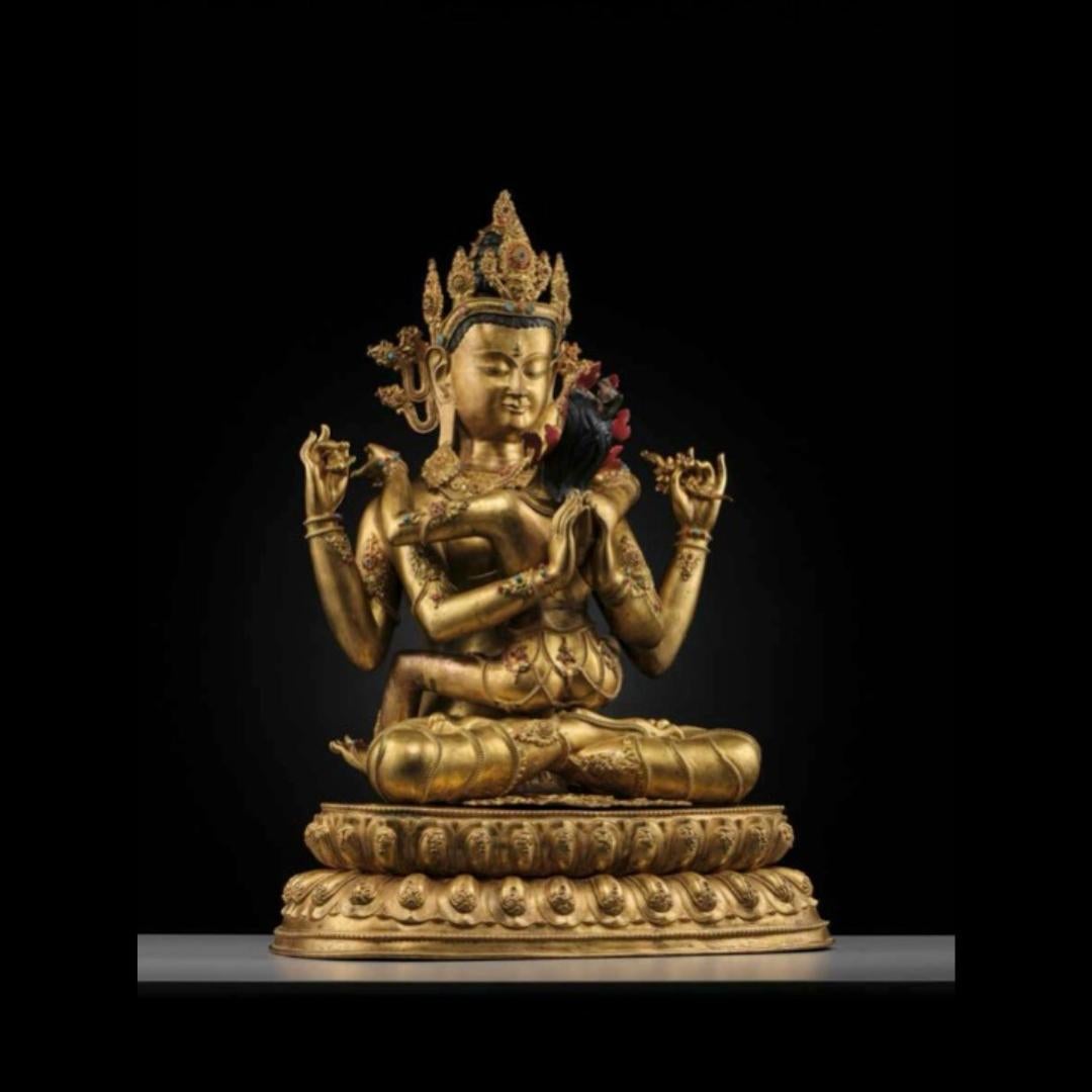 Finement moulée, la divinité est assise en dhyanasana sur un double trône de lotus aux bords perlés, en union (yab yum) avec sa consort Shakti dont les jambes s'enroulent doucement derrière elle. Les bras principaux de la divinité sont tenus en