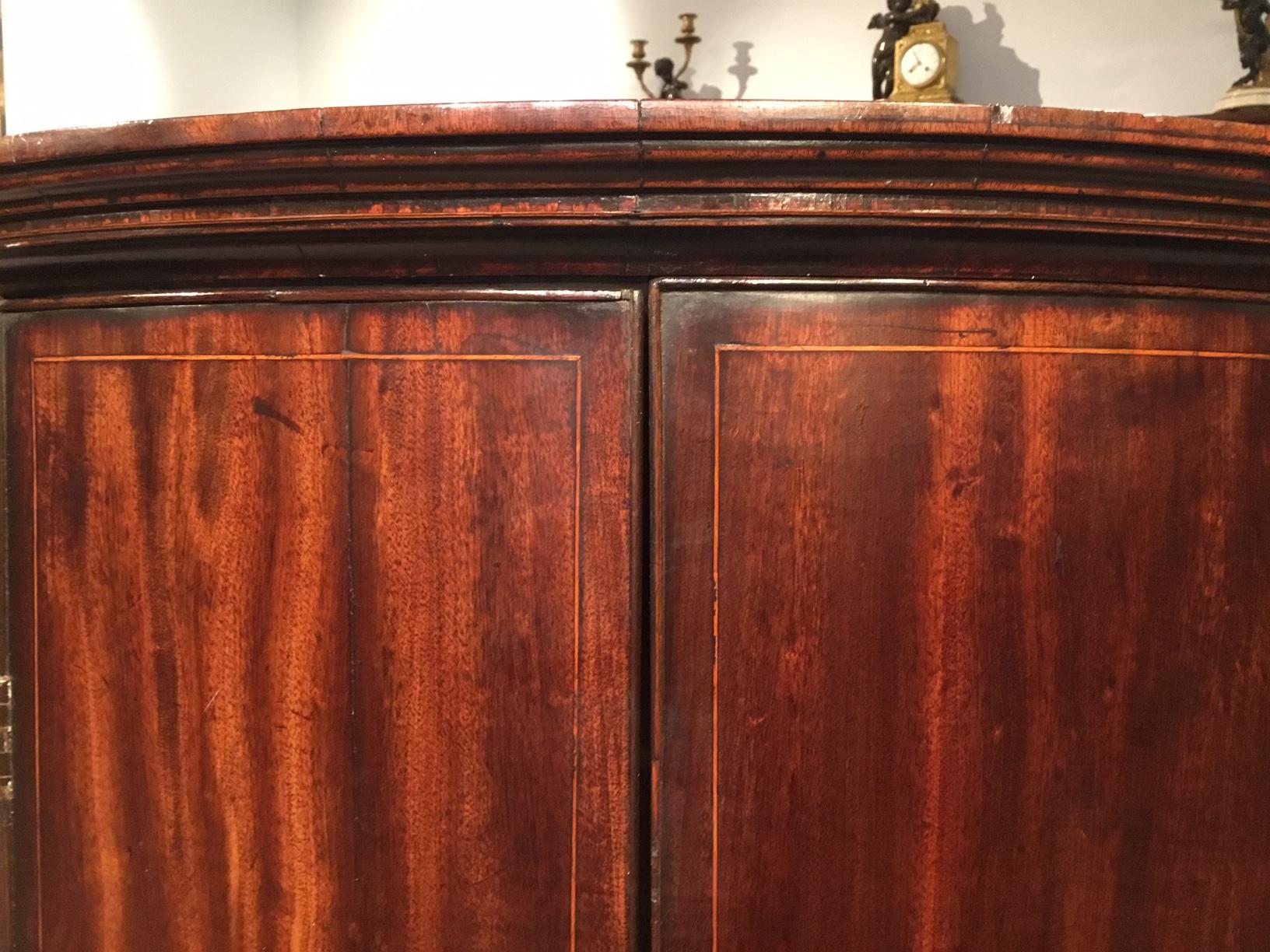 A mahogany George III Period bow front corner cupboard. Having a moulded cornice above twin solid mahogany doors with line inlaid details, opening to reveal a painted shelved interior. English circa 1780-1800

Dimensions: 28