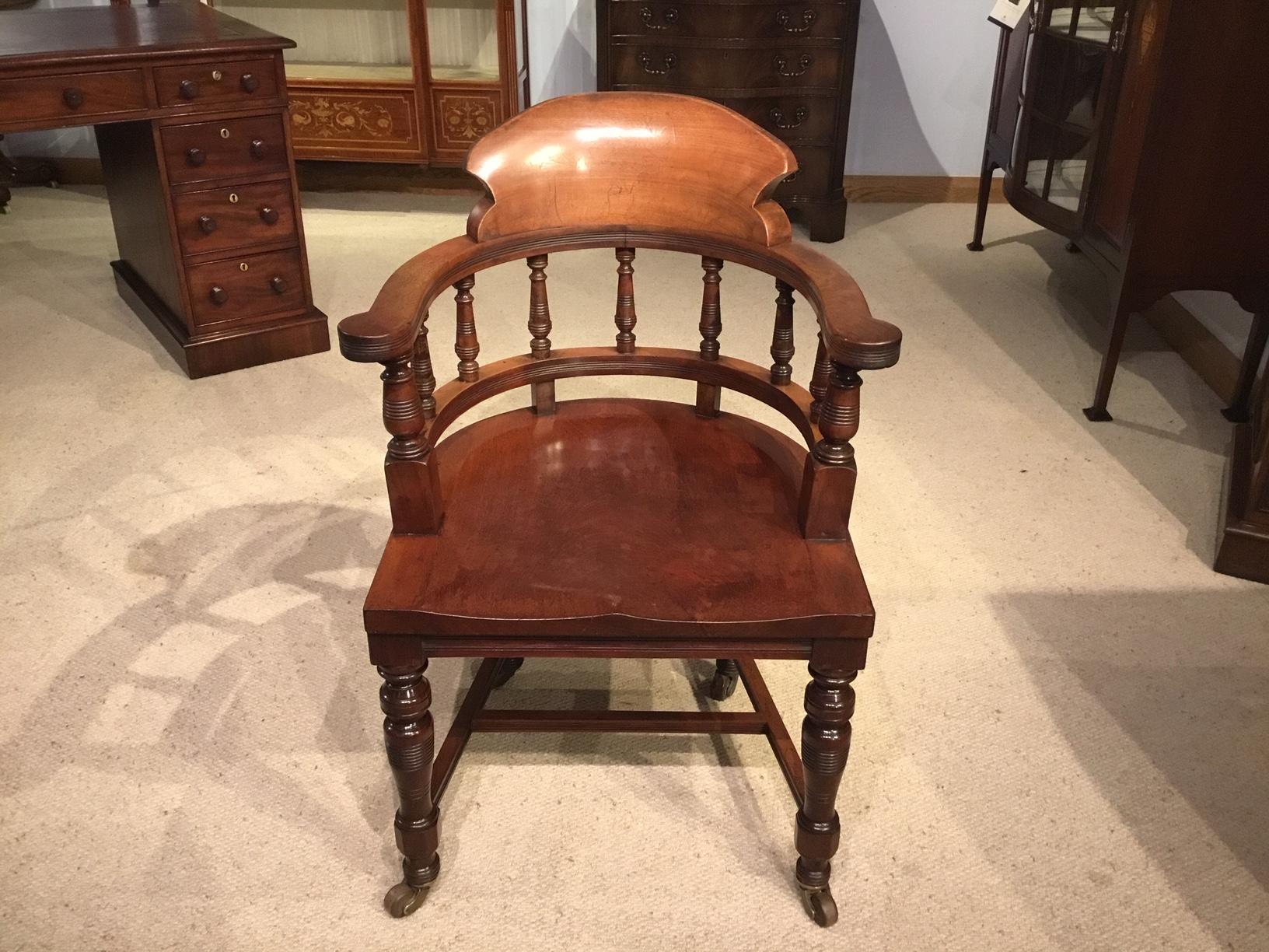A Late Victorian Period mahogany desk armchair. Having a curved solid mahogany back support, open arms and turned spindle supports. Having a generous solid mahogany dished saddle seat. Standing on ring turned supports with brass and porcelain