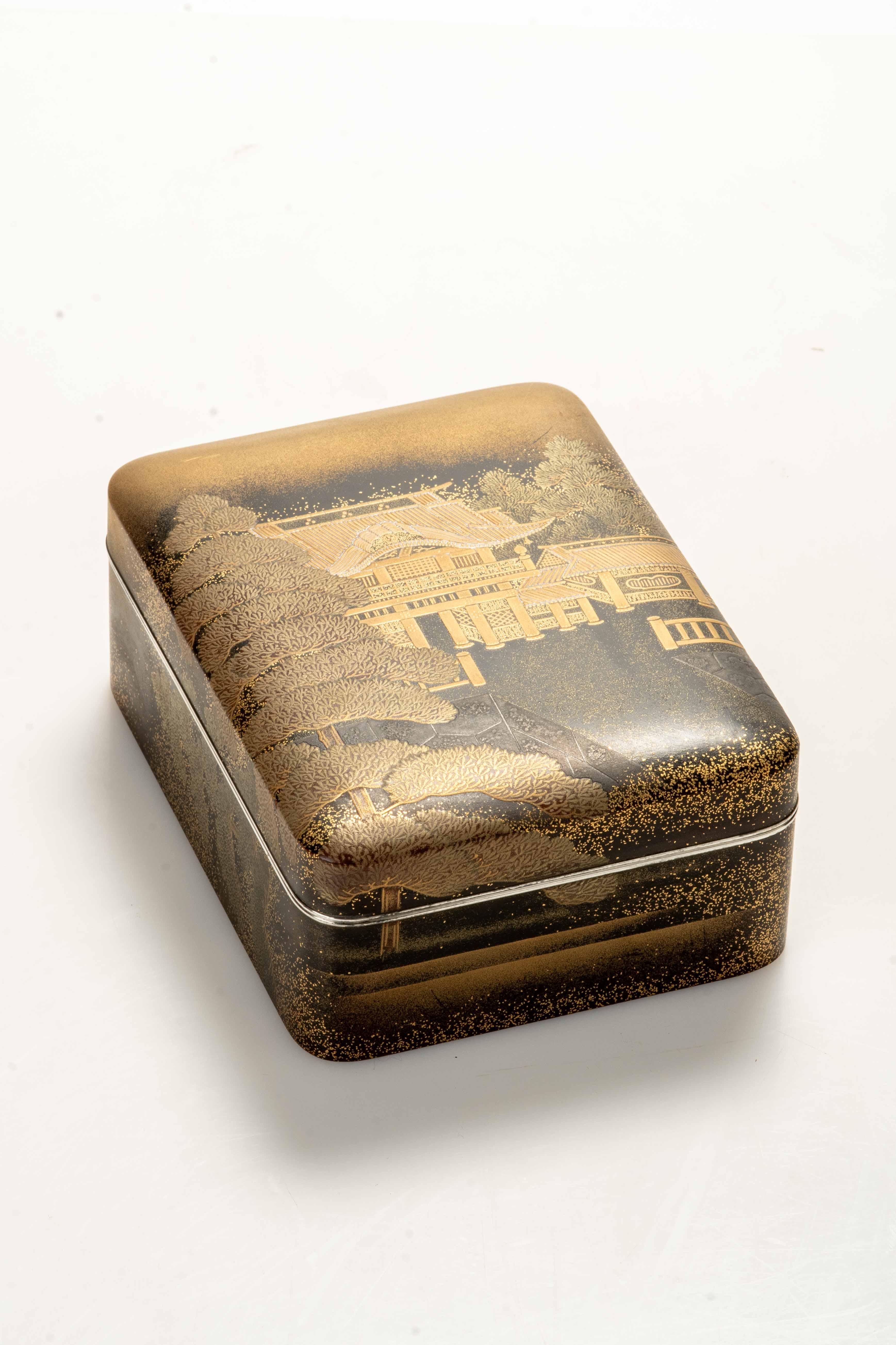 Caja tebako de laca maki-e embellecida con adornos dorados que representa un templo budista rodeado de majestuosos pinos de Matsu. El interior y la base están acabados en laca nashiji. Los bordes de la caja y la tapa están elegantemente acabados en