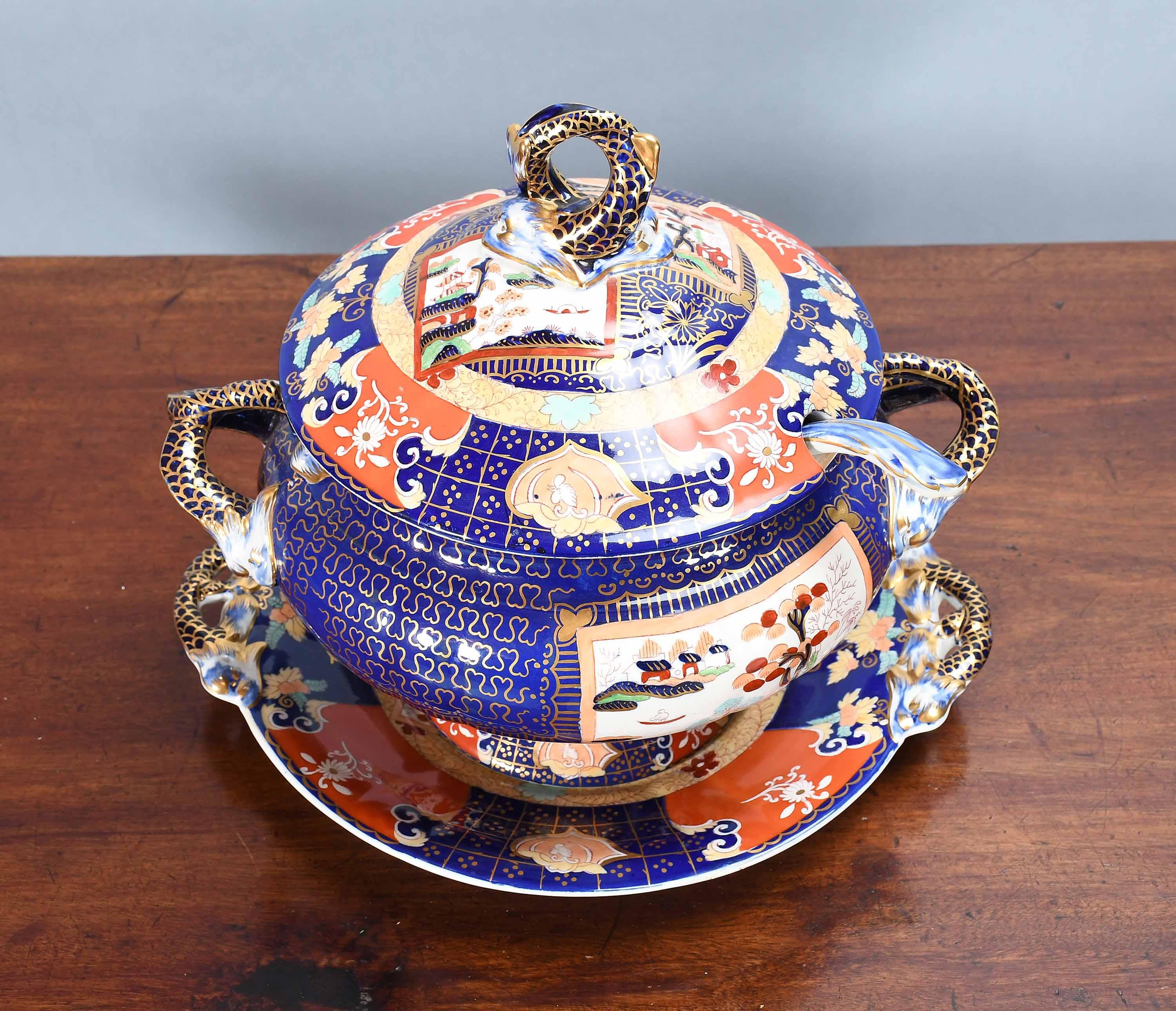 Eine Suppenterrine aus Eisenstein von Mason mit Deckel und Ständer mit doppeltem Landschaftsmuster. In leuchtenden Blau-, Gold- und Rottönen. Mit Delphingriffen an der Terrine, am Ständer und am oberen Rand des Deckels. Die Schöpfkelle ist repariert