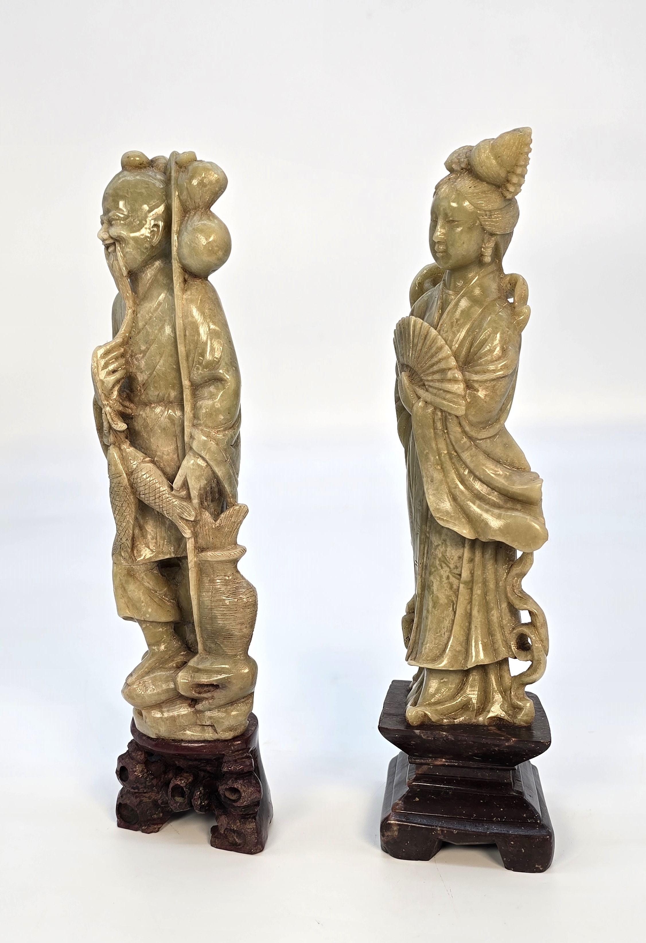 This decorative pair of soapstone carvings depicts traditionally dressed figures of a man and woman. The woman is dressed in ornate robes, and she is holding a fan. The man is a fisherman, dressed in simple robes, and he is carrying his catch of