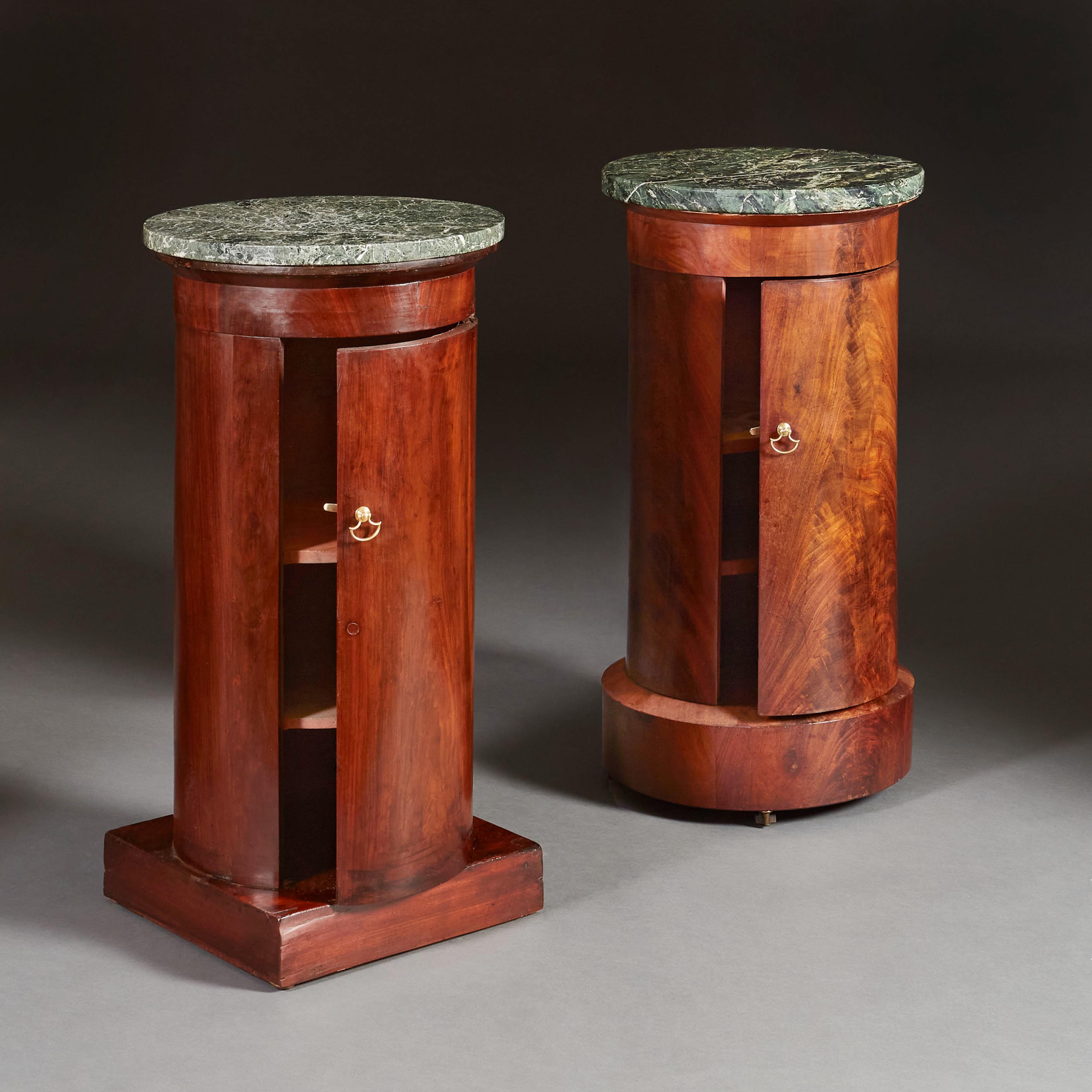 A matched pair of Empire mahogany pedestal cupboards of cylindrical form, both with green marble tops, and two shelves to the interior. One with circular plinth base and castors, one with square plinth base. With Brass handles.

Circular base