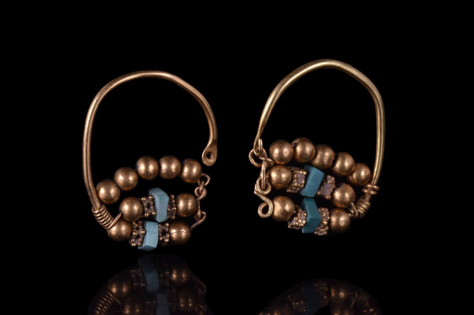 A matched pair of gold earrings, each made of a wire elongated hoop culminating with a teardrop turquoise cabochon. The bottom half of the loop is decorated with three rows of gold baubles, and the turquoise adornments are flanked by four beaded,