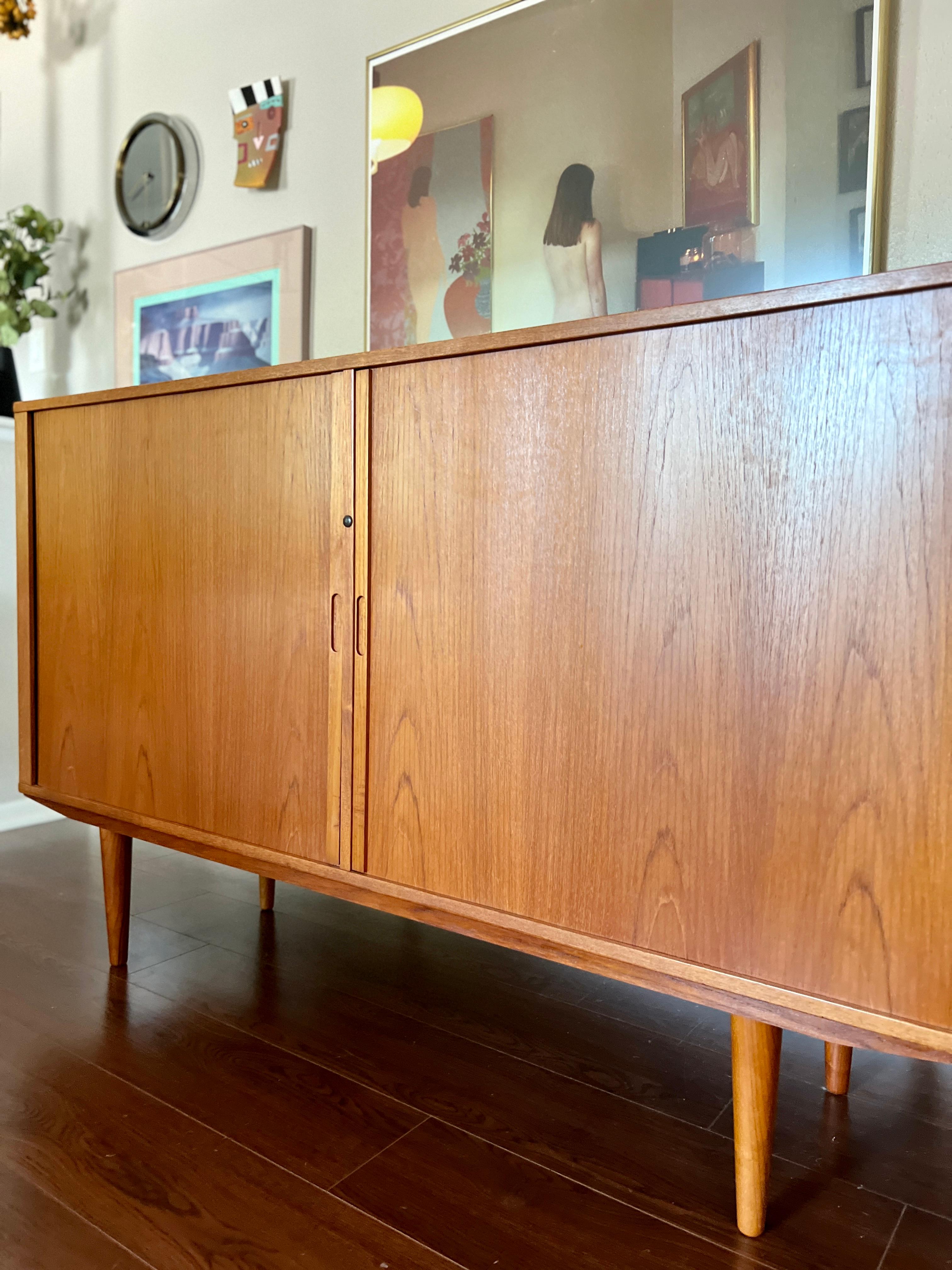 A MCM minimalist teak sideboard featuring disappearing tambour doors and tapered legs. This classic Danish modern storage piece has a beautiful contrasting birch interior with three adjustable shelves. Maker is unknown. This piece is in very good