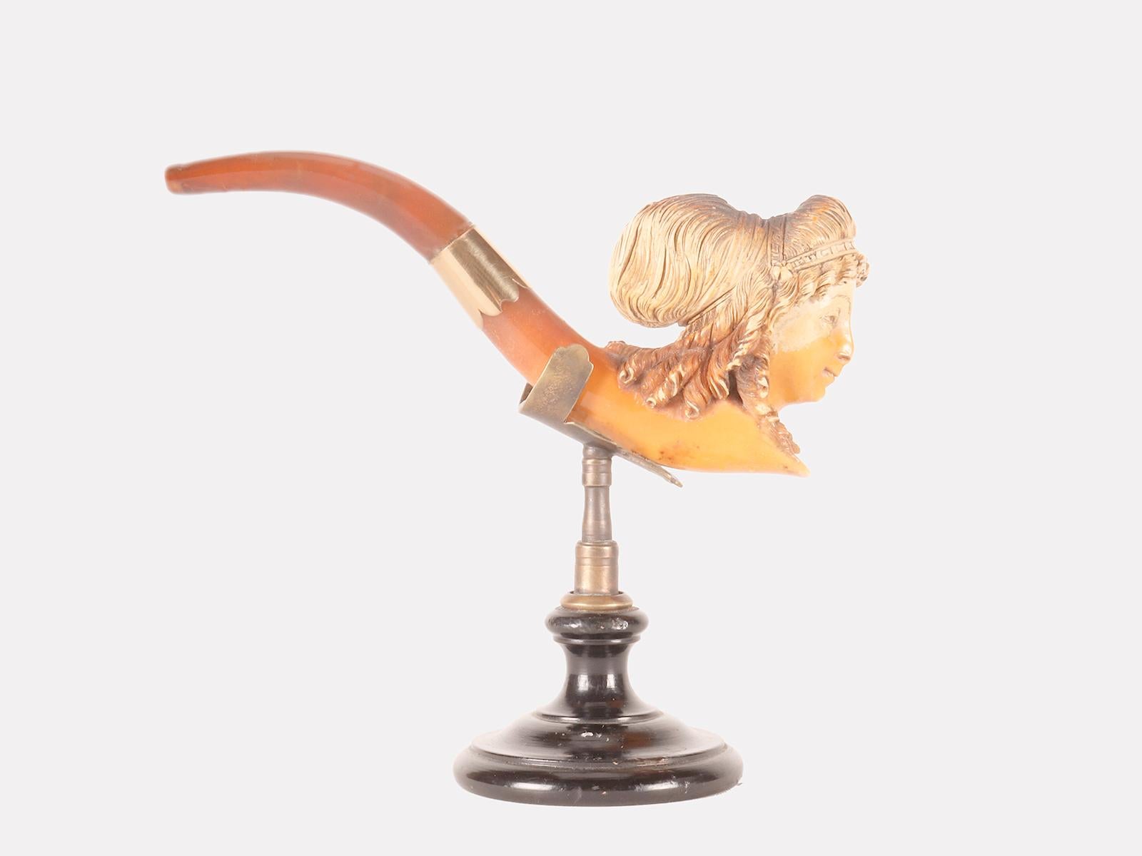 Pipe en écume sculptée, avec embouchure en ambre et bandeau en or. Dans le fourneau de la pipe, une tête de femme est représentée avec des cheveux rassemblés par une coiffure de rubans et de fleurs. Vienne, Autriche vers 1890. (Le socle est destiné