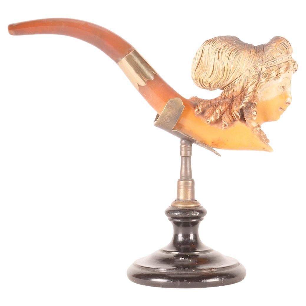 Carved meerschaum pipe, with amber mouthpiece and gold band. In the bowl of the pipe, a woman's head is depicted with hair gathered with a hairstyle of ribbons and flowers. Vienna, Austria circa 1890. (The stand base is for photographic use only,