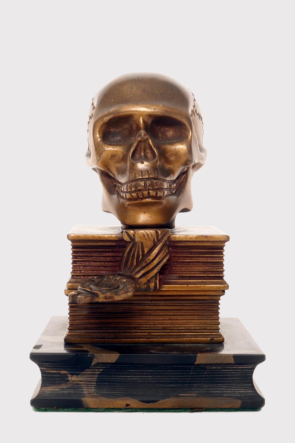 Una scultura Art Decò che raffigura un Memento Mori. Alla base della scultura troviamo un libro chiuso, realizzato in marmo Portoro, sopra il quale si trovano due libri chiusi in bronzo che sostengono la scultura di un teschio, anch'essa in bronzo.