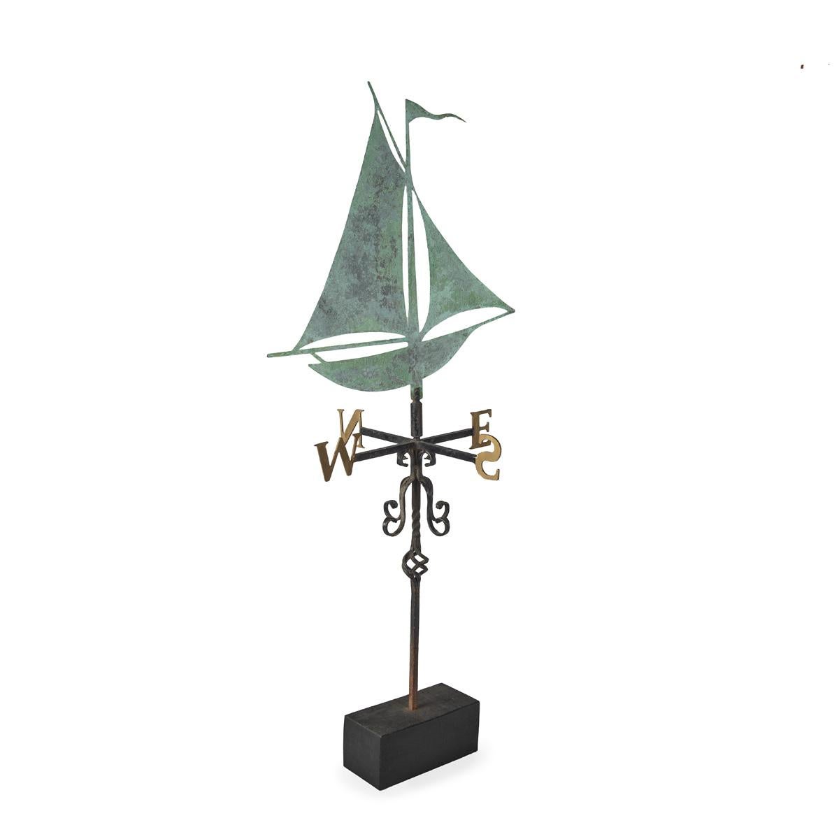 A metal weathervane by Martin Scorey, comprising a sailing yacht set above a four arms with the four compass points N, E, S and W above scrolls, set into a rectangular  wooden base..  English, circa 2010.


