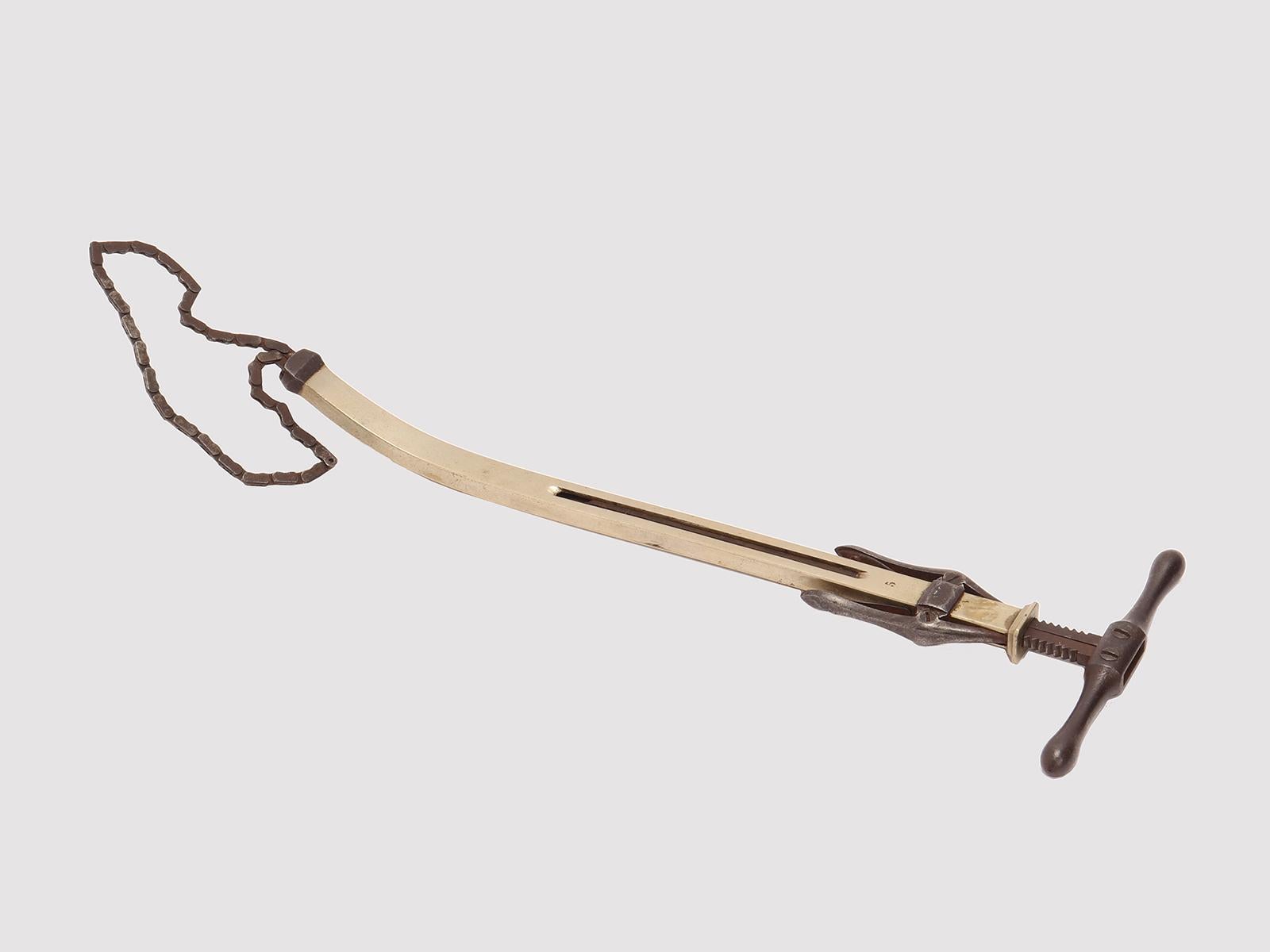 A 19th-century surgical instrument. An amputation chain saw with a white metal body and iron stops, extractor, and chain. The bar-shaped body has a rectangular section that curves slightly at the end. Inside runs the chain pulled and released by