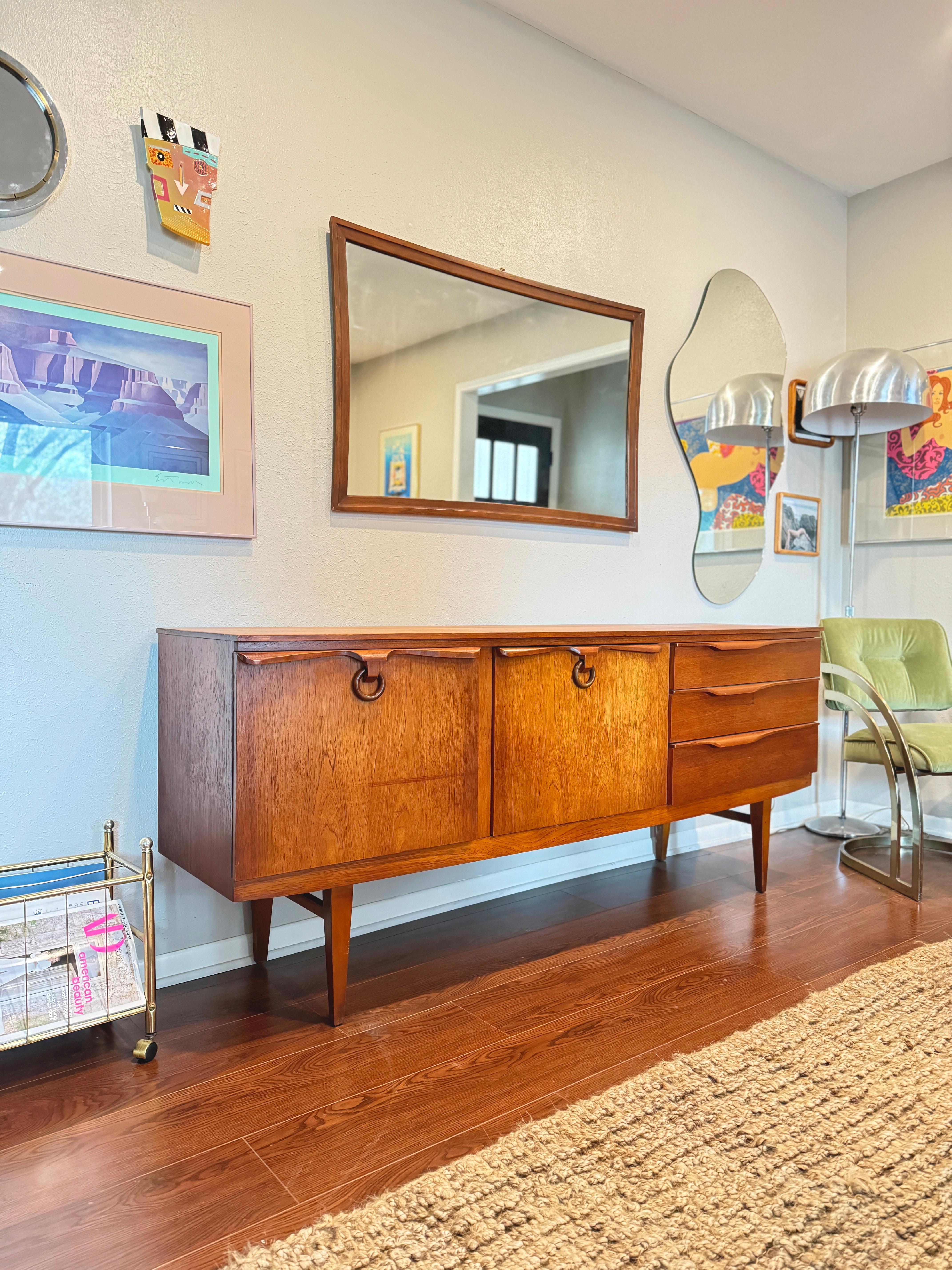 A mid century modern teak sideboard by Beautility, circa 1960s. This lovely sideboard features three drawers, one swinging door with two shelves, and a drop down door. With its sculptural elements, from the wooden ring pulls, tapered legs, and