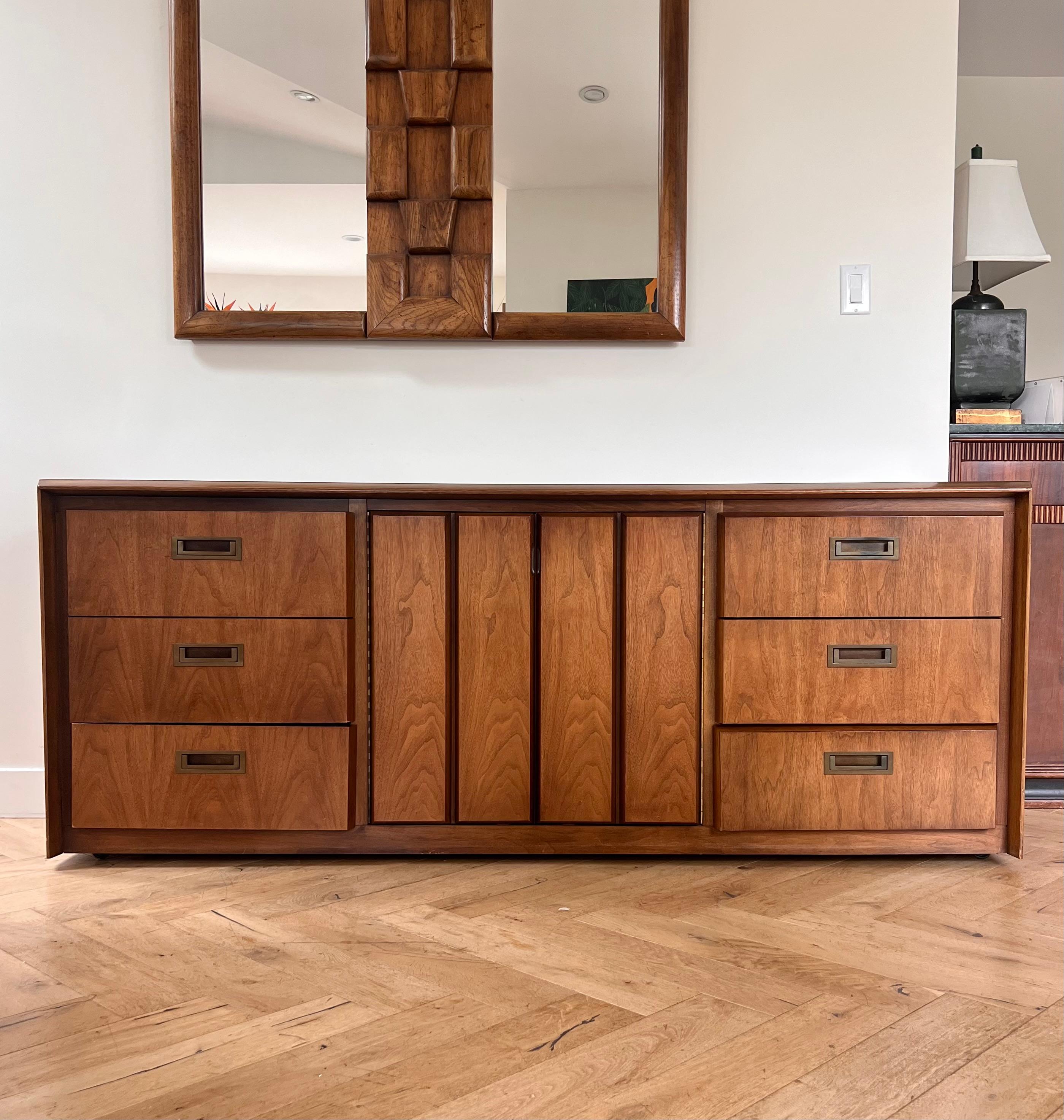 A stunning mid century modern dresser, sideboard, or buffet in rich walnut wood by American of Martinsville, circa 1960. Featuring brutalist geometry, in the angled accents of the drawers, and original hand-forged metal hardware, this piece is at