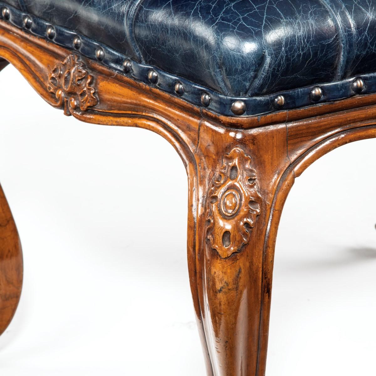 A mid-Victorian walnut stool, the shaped rectangular seat reupholstered in deep-buttoned distressed blue leather, raised on cabriole legs with flowers carved on the knees. English, circa 1870.
  
