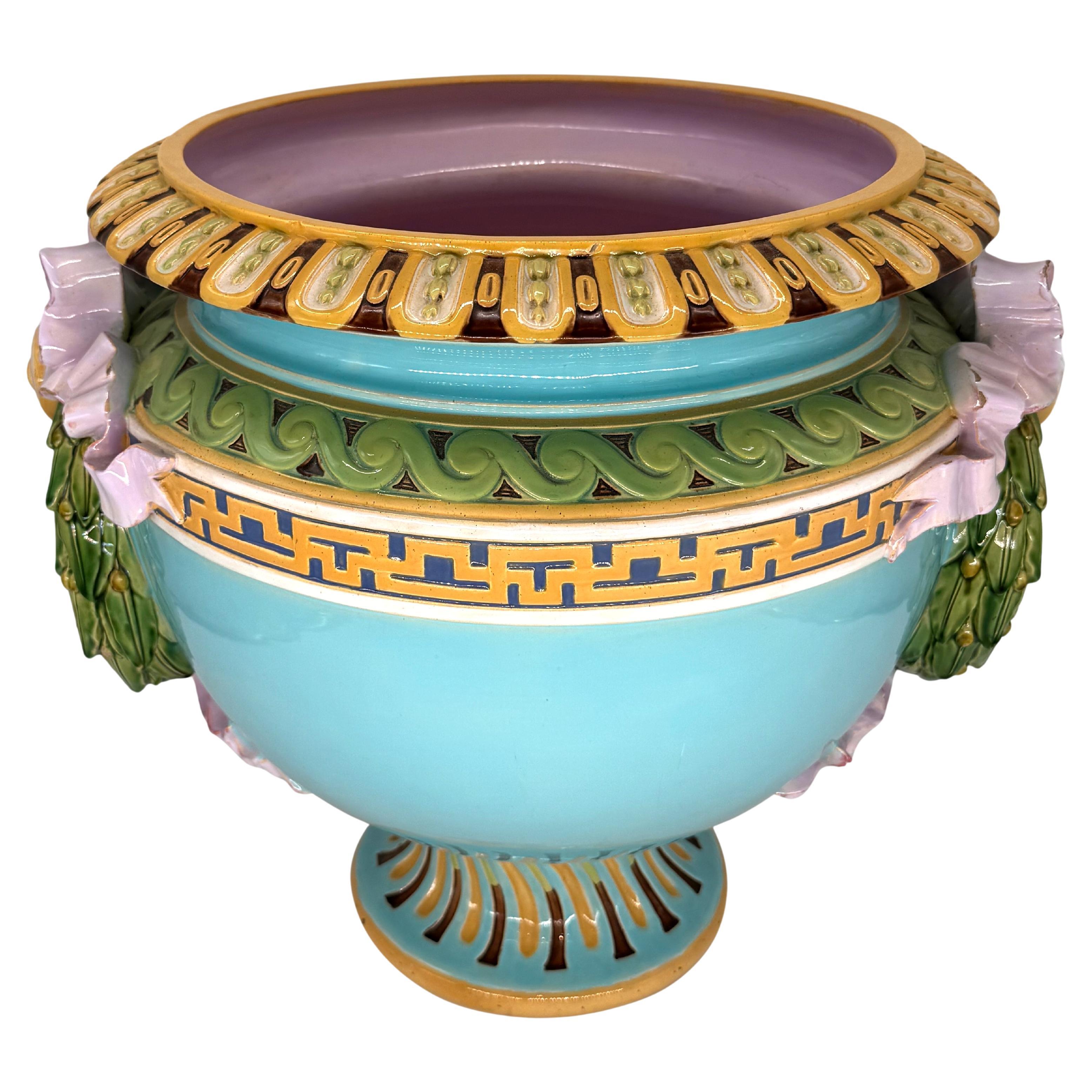 Jardinera de mayólica Minton, cuerpo en forma de urna con asas aplicadas en forma de corona de laurel atadas con lazos rosas sobre fondo turquesa, hombro y borde moldeados con bordes neoclásicos, interior esmaltado en rosa, sobre base con patas,