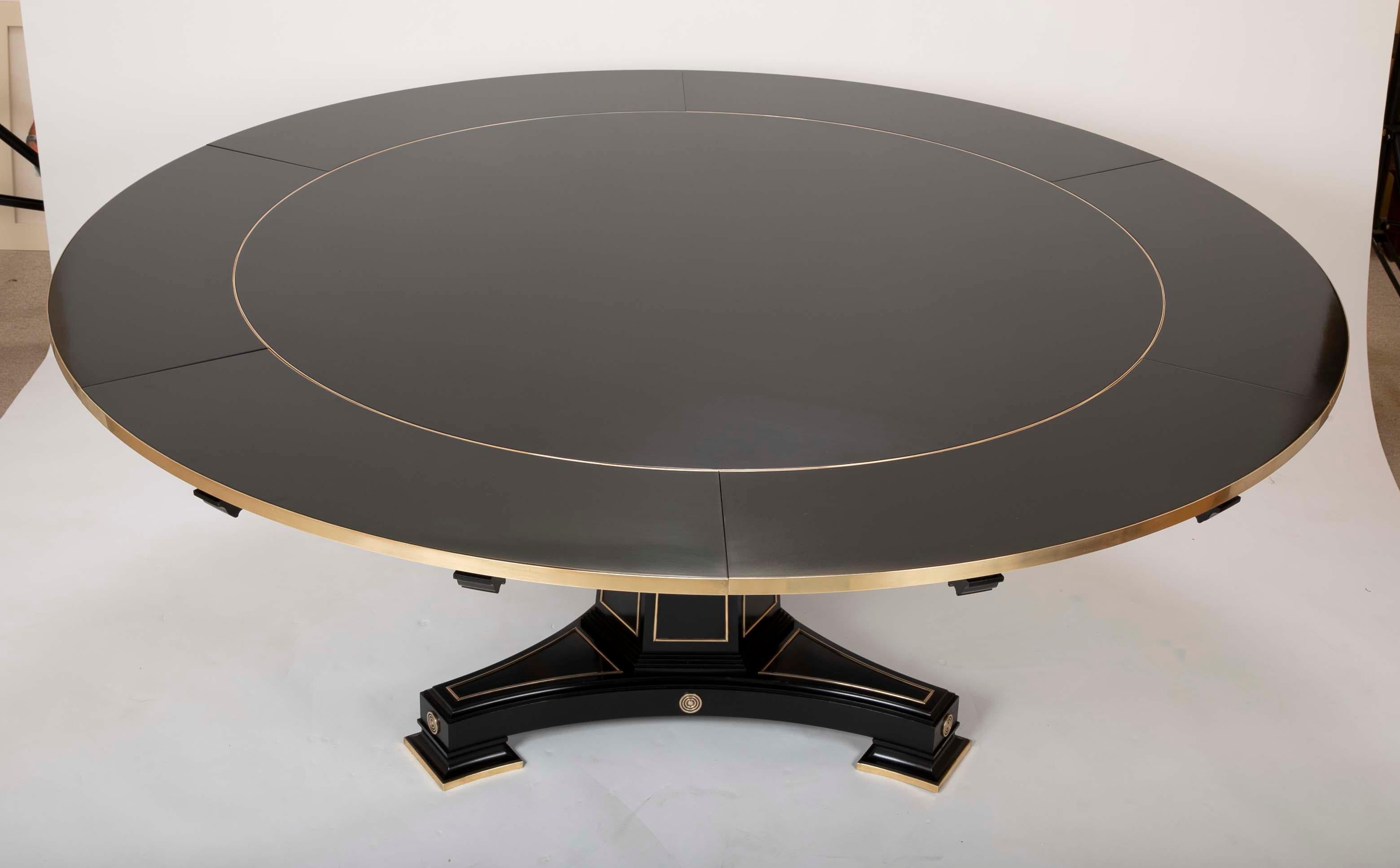 A modern ebonized expandable dining table in the Empire style. Designed by David Linley, The table has a central brass banded round top that accepts 6 brass banded leafs raised over a central tripod pedestal. There are also tracery brass decorations