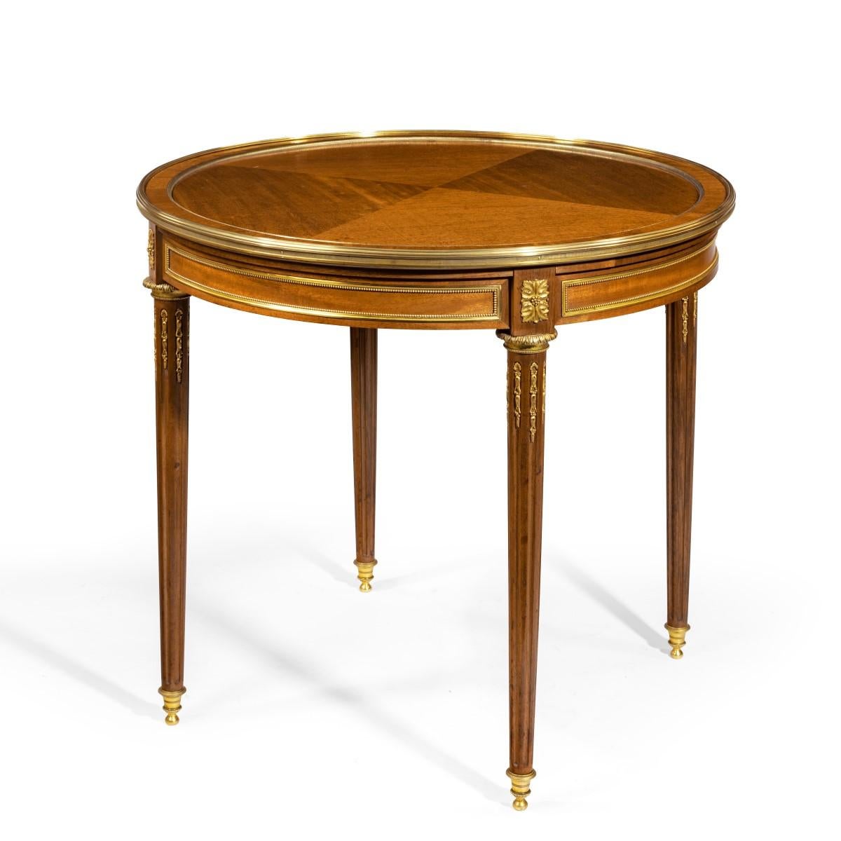 A Napoleon III mahogany side table, the circular top with two disguised frieze drawers alternating with two slides, set upon stop-fluted tapering legs on turned brass castors, decorated with quarter veneers and ormolu mounts, French, circa