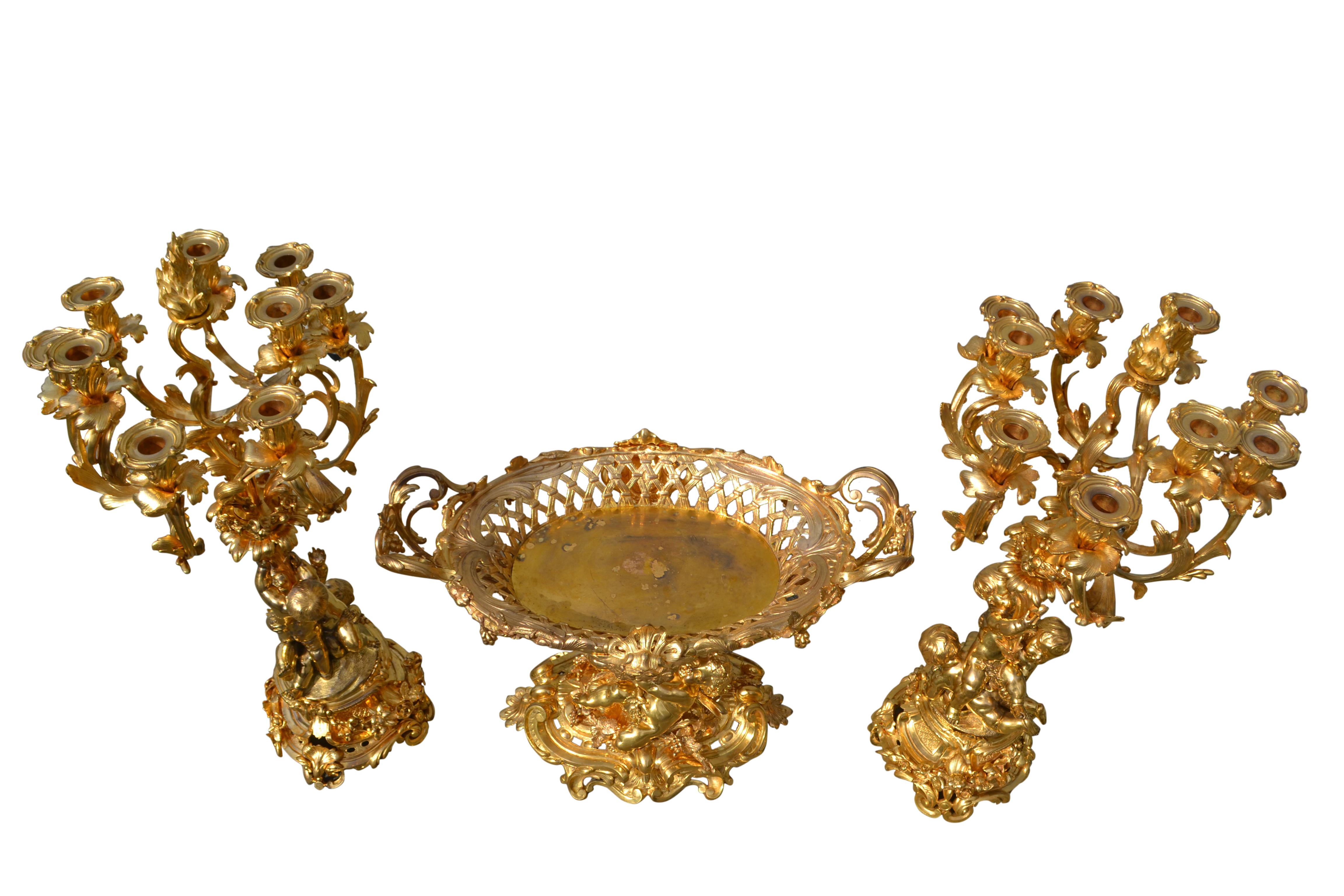 The candelabra feature three putti standing on an elaborate rocaille base holding aloft three flower and vine encrusted scrolling branches each with three candle nozzles 

The large gilded bronze table centre is supported by two winged putti