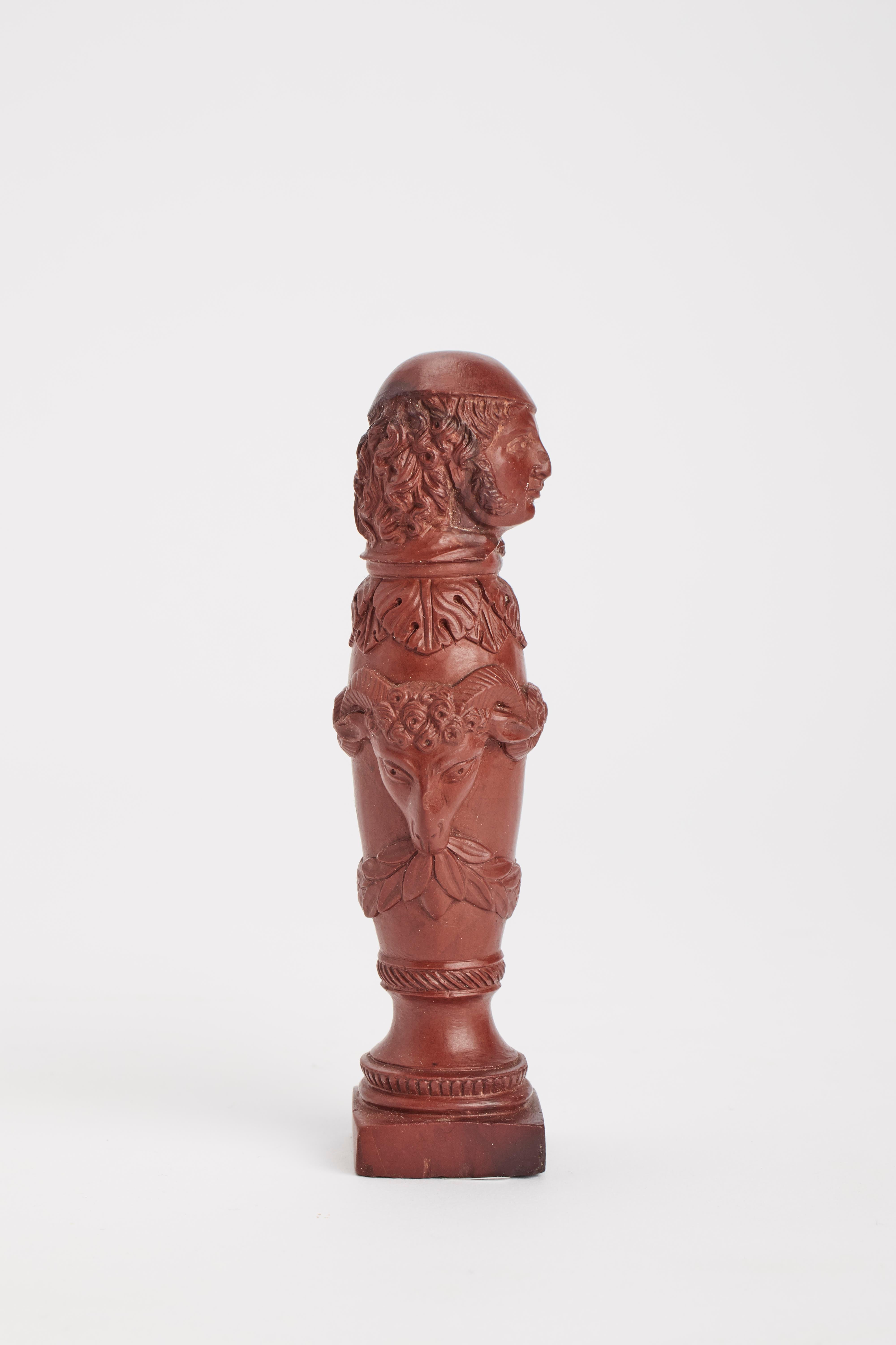 A Neoclassical Grand Tour seal, carved out of red lava, depicting an Italian character with beret, garlands and ram heads over a stylized column. Italy 1840 ca.