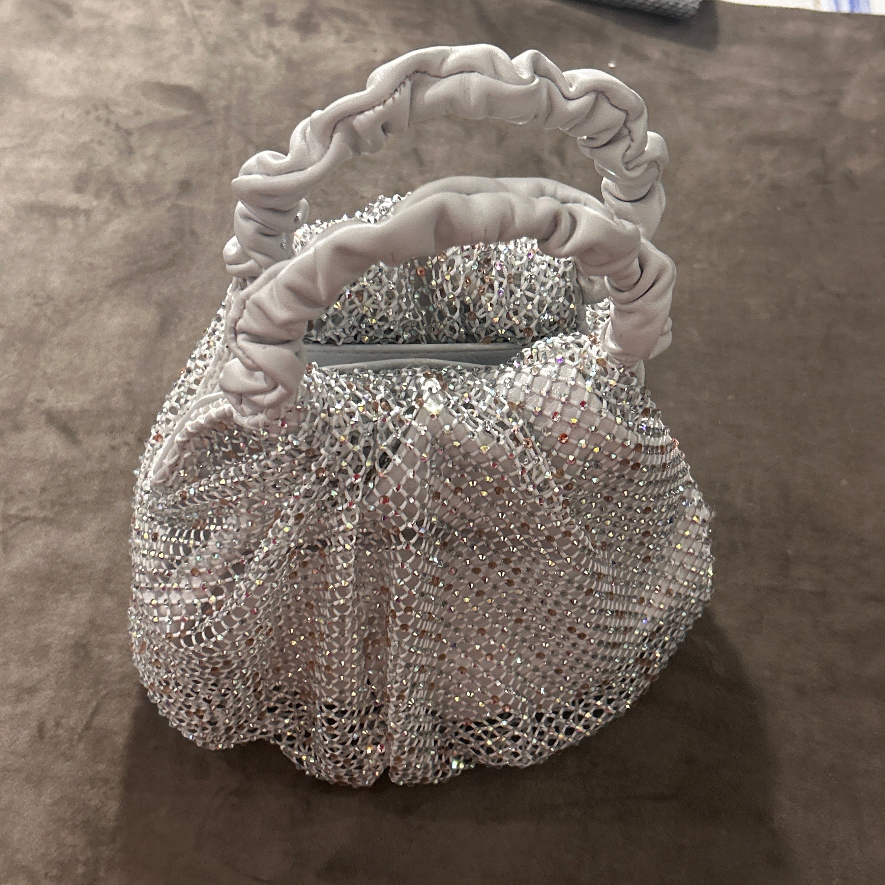 This Gedebe handbag embodies a blend of glamour and playful sophistication, showcasing the brand’s flair for combining luxe materials with distinctive design. The bag features a softly structured, ruched handle crafted from white leather, giving it