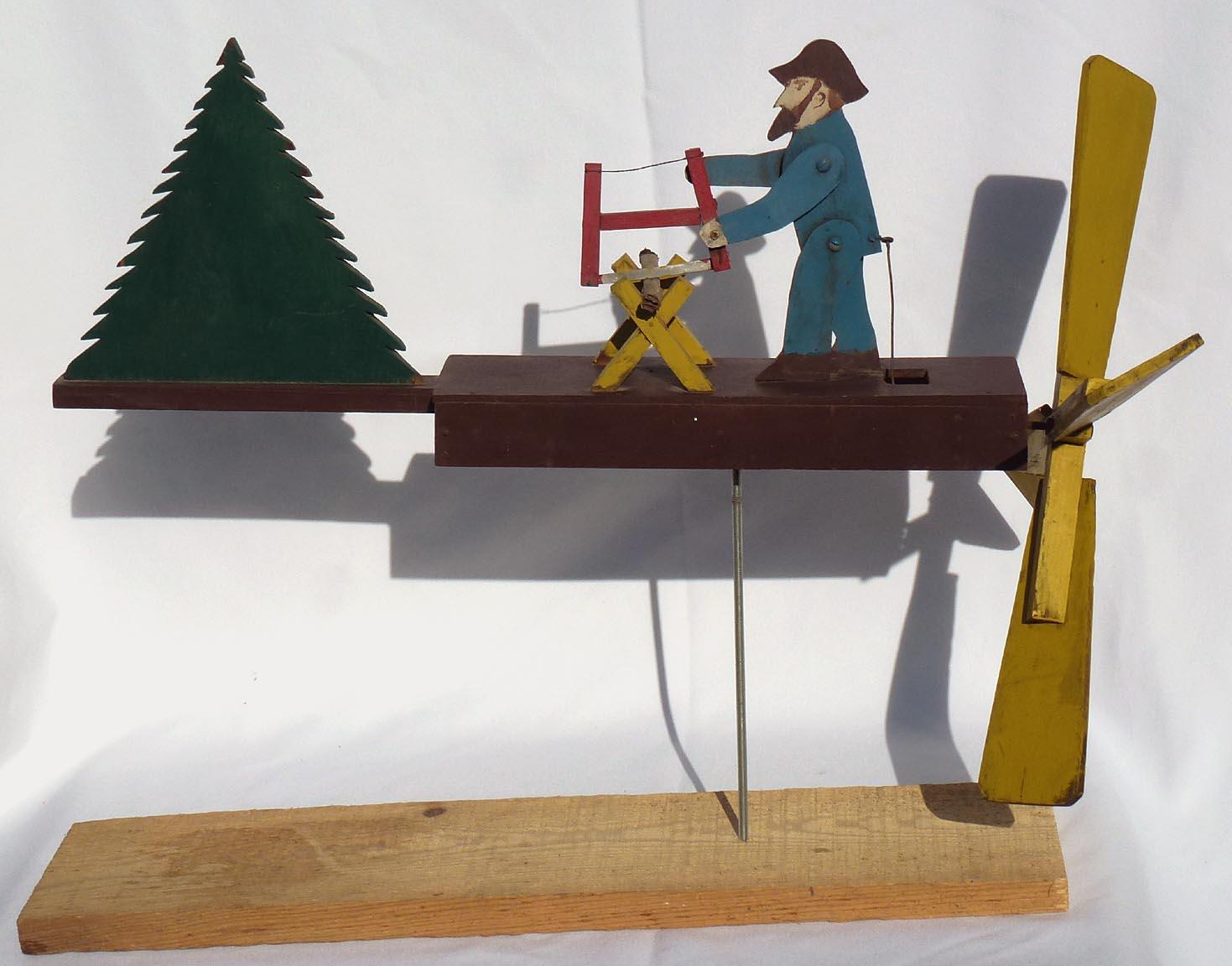 This is a nicely painted whirligig of a bearded man sawing wood. It is made of wood with four yellow blades and a pine tree for a tail. From the second half of the 20th c., it is in very good, weathered condition. It has been mounted on a newer wood