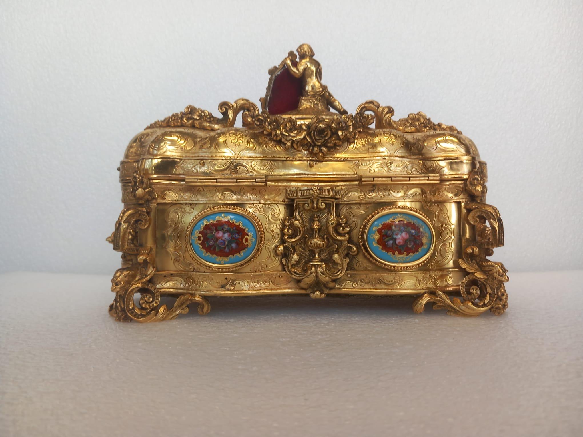 A nineteenth century French ormolu casket, in the medieval style, with a cherub sitting on top of the hinged lid. 
The body has 4 hand painted cartouches of flowers at the front.

The gilded ormolu is etched with scrolls and floral designs whilst