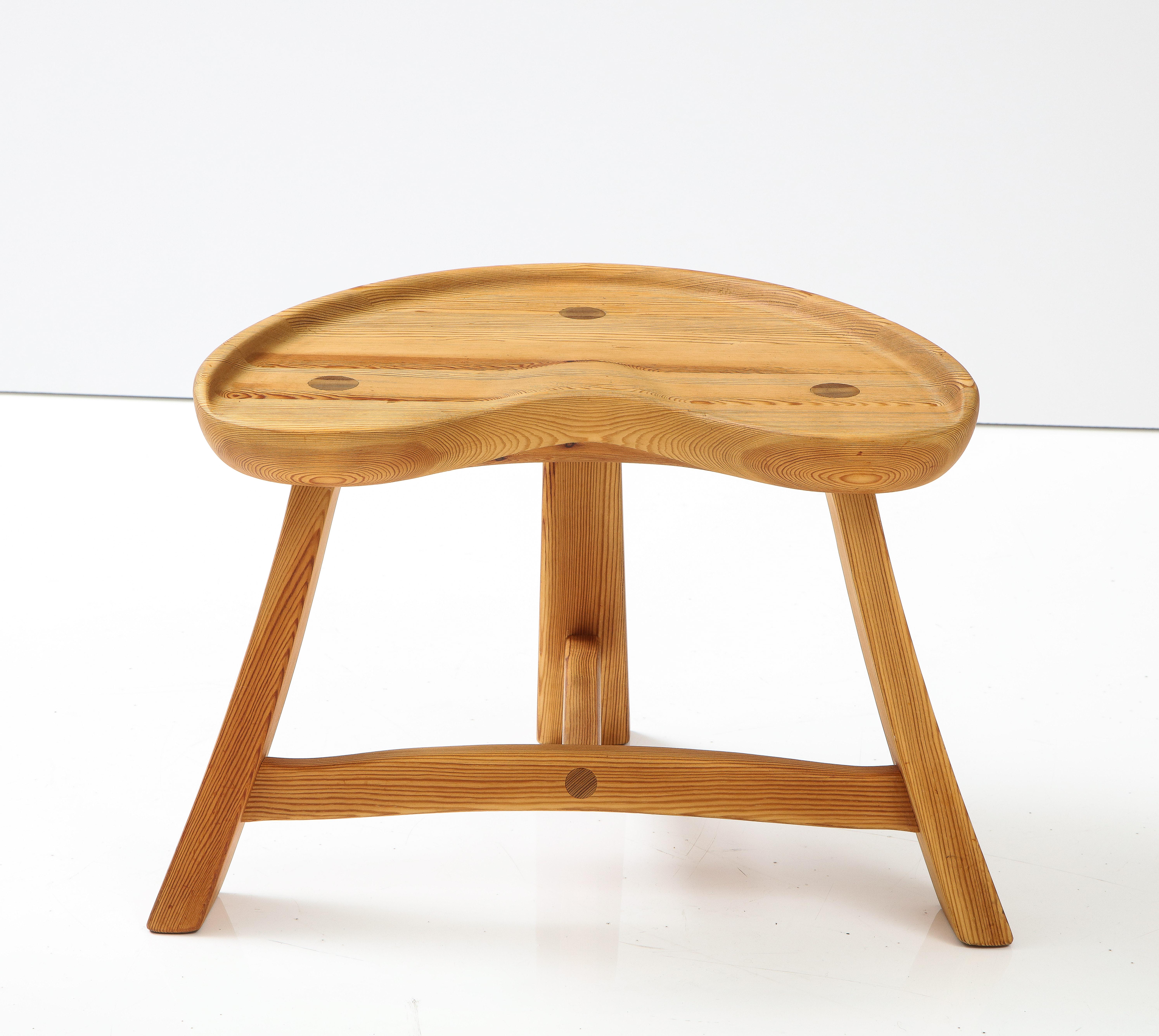 A carved solid pine three leg stool by Krogenæs Møbler, Norway, Circa 1950s-60, with a carved tractor form seat raised on three rectangular legs joined by a T-form stretcher. A truly sculptural piece of seat furniture.