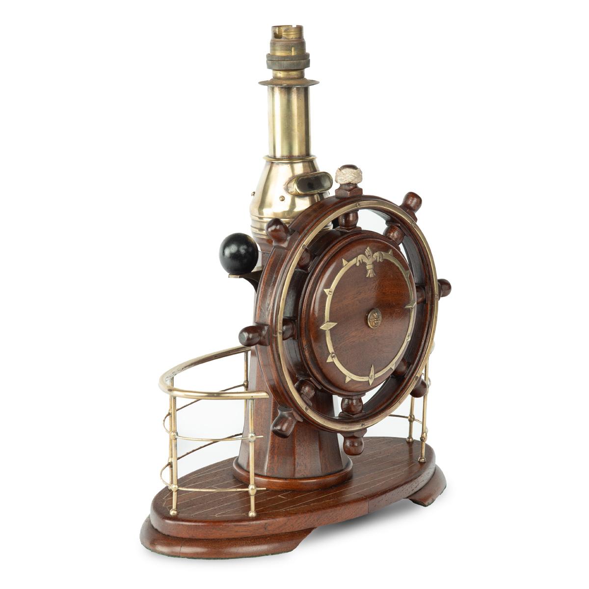A novelty maritime teak, mahogany and brass table lamp, comprising a steering wheel attached to a binnacle, all within a brass railing, the wheel inlaid with a brass circlet and stylized fleur de lys, with the central button embossed with a naval
