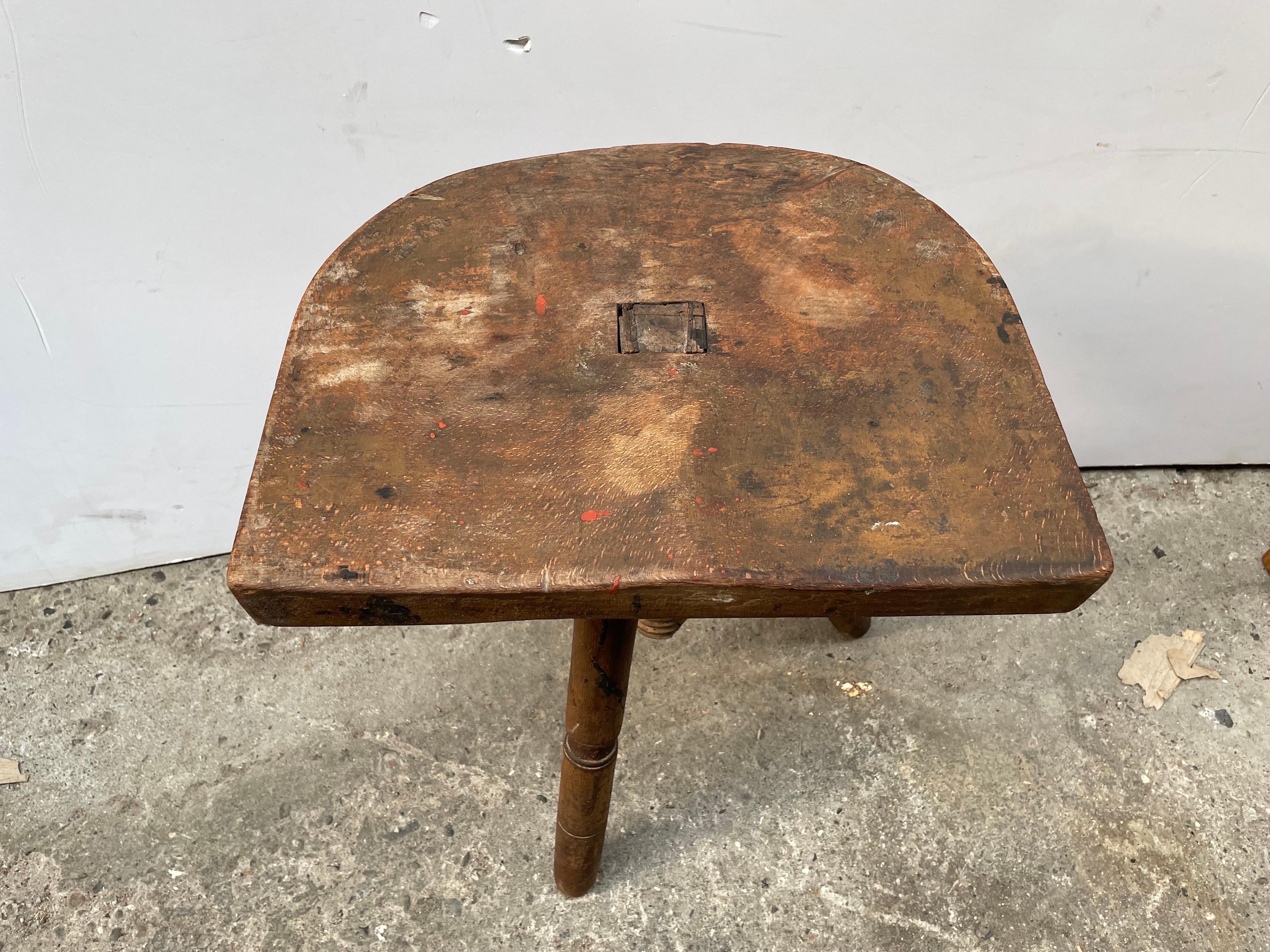 An old stool with a carved seat raised over 3 legs. One of the legs have been replaced with a piece of wood. It is truly one of a kind piece of antique furniture.