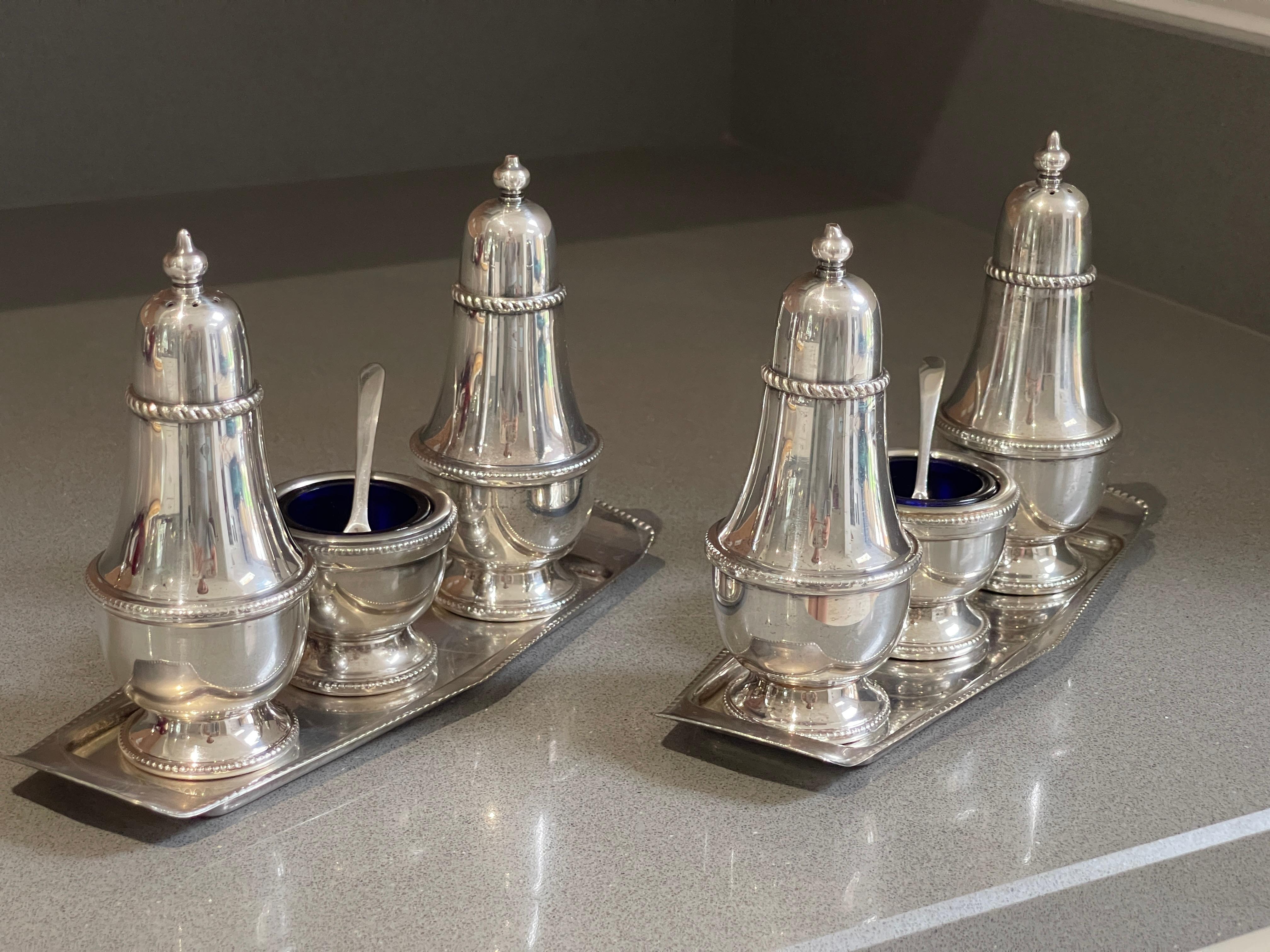 A pair of modernist Silver Condiment sets comprising a Salt shaker, a Pepper Shaker, a Mustard Pot & a spoon. The mustard pot has a blue glass bowl and a silver spoon. Each item's exterior has an elegant tress pattern around it.

Made in London in