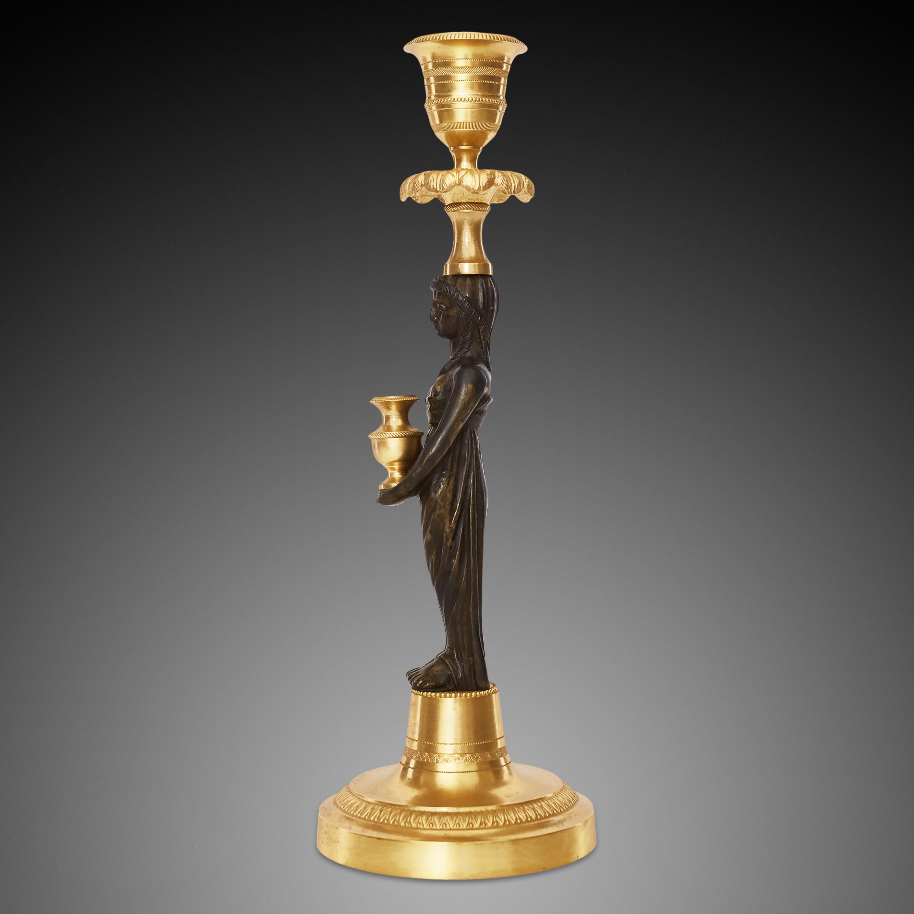 A pair of gilded bronze patinated candlesticks, each depicting a figure of a woman holding a golden-colored jug in her hand. The candlestick at the top is decorated with a pattern resembling a crown of leaves.