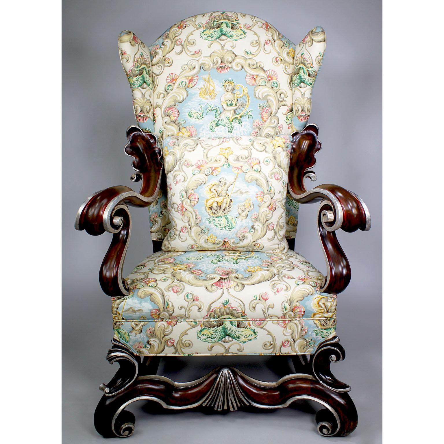 A large nicely carved pair of Baroque Revival style Walnut and Parcel-Silver Leaf Winged Throne Armchairs. The tall padded winged back armchairs, with scrolled carved walnut frames in a dark finish stain, each with finely carved open-armrests with