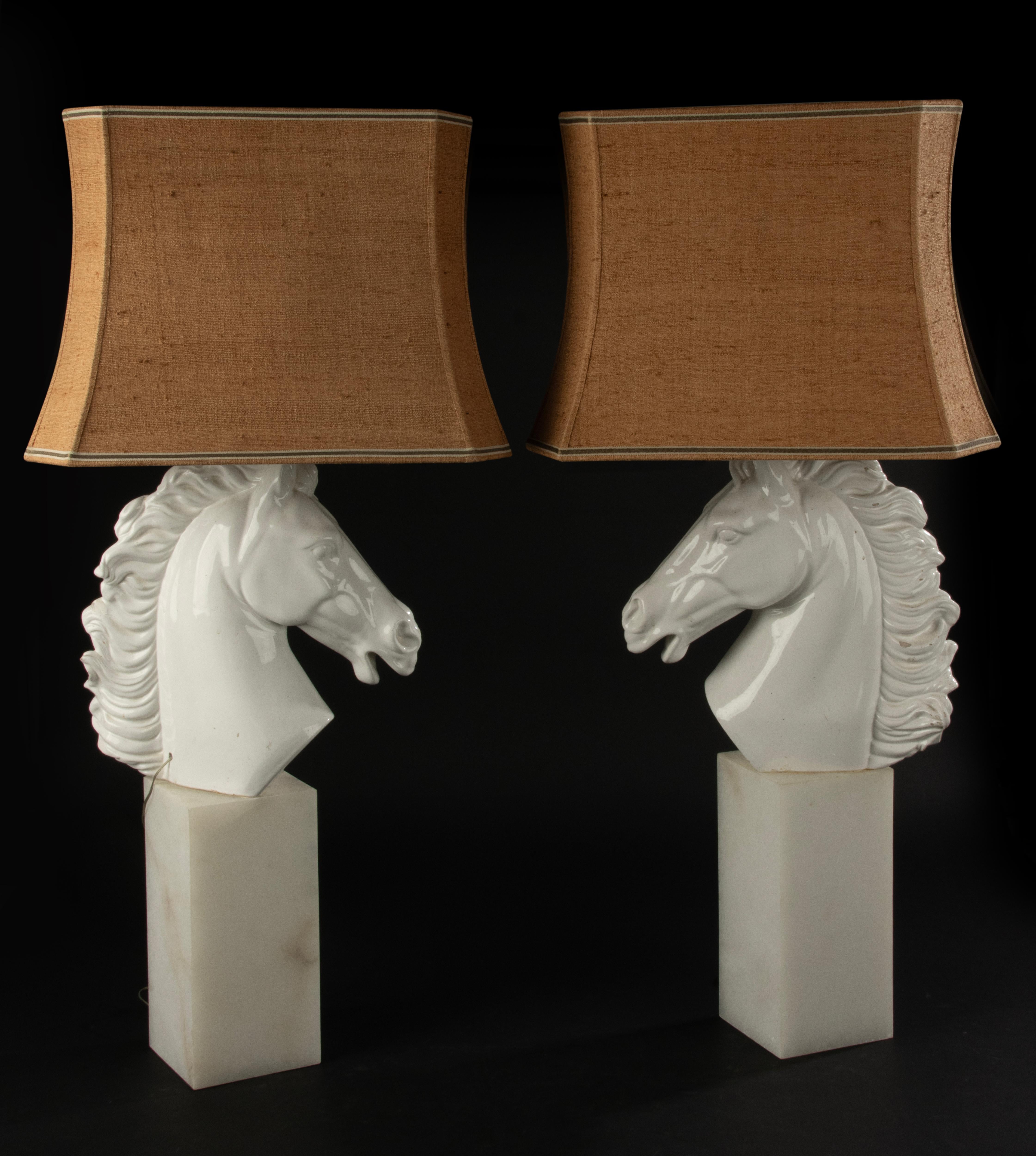 Una straordinaria coppia di grandi lampade da tavolo italiane Hollywood Regency con eleganti teste di cavallo in ceramica bianca montate su basi in marmo massiccio. Questi imponenti pezzi unici sono completi delle loro tonalità di tessuto originali,
