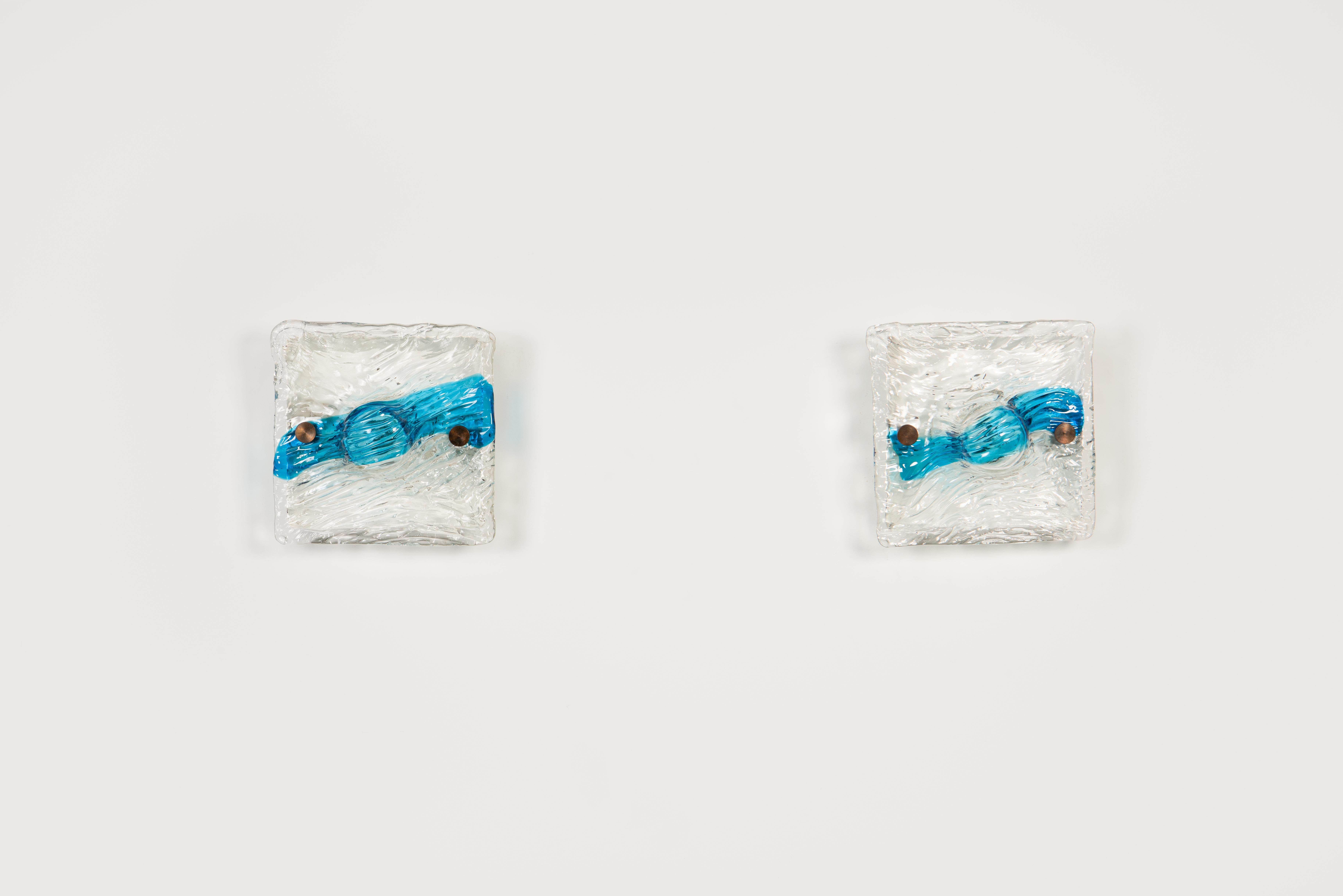 Pareja de apliques de cristal de Murano cuadrado soplado a mano, transparente y azul, con pantalla abombada en el centro. Soporte de pared cuadrado de metal lacado en blanco. El cristal se sujeta con dos tornillos redondos ornamentales de latón. Las