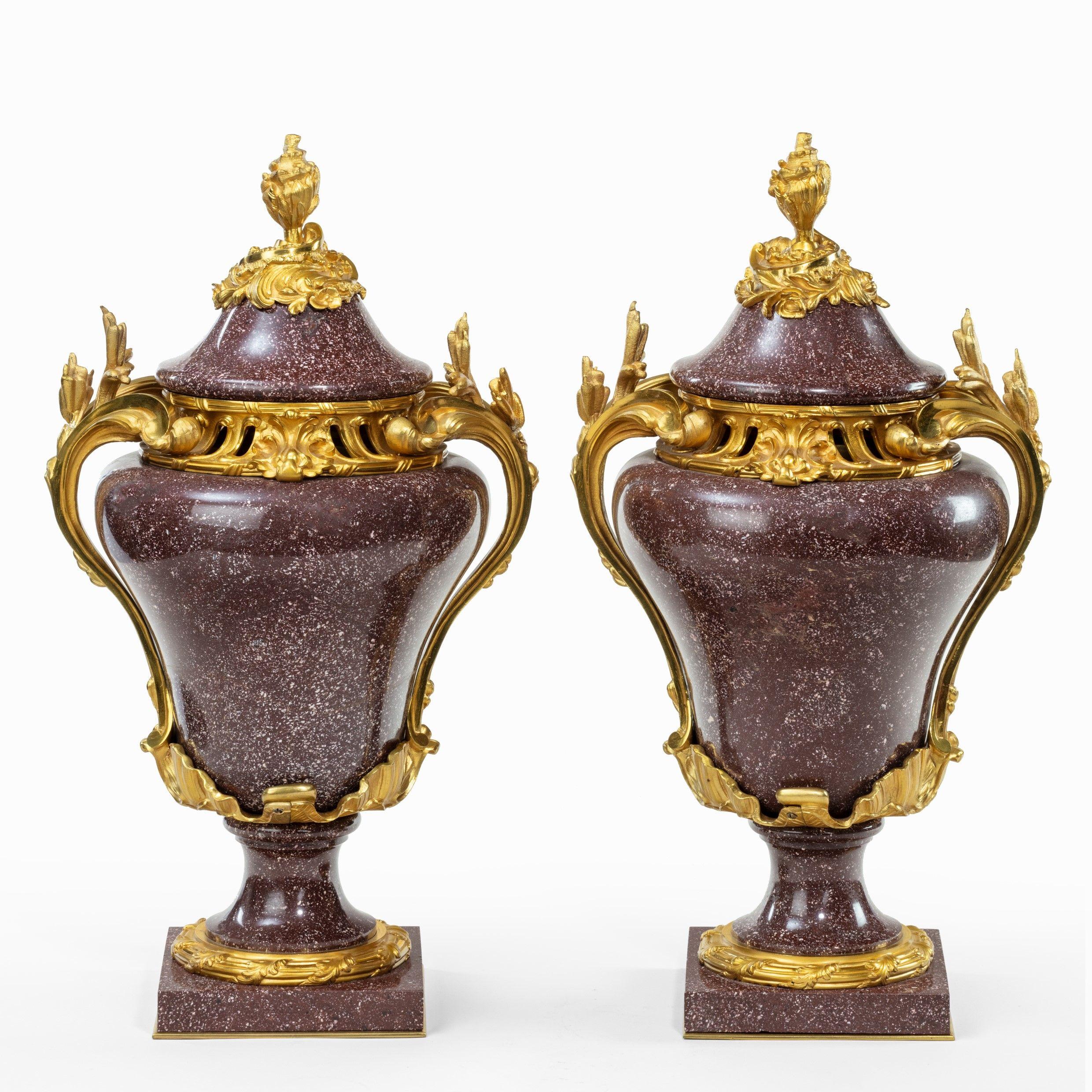 A pair Napoleon III Egyptian imperial porphyry urns and covers, each with a shaped cover and baluster body on a concave support and square base, applied with high quality ormolu mounts comprising a flower-bud finial, arched foliate handles, an