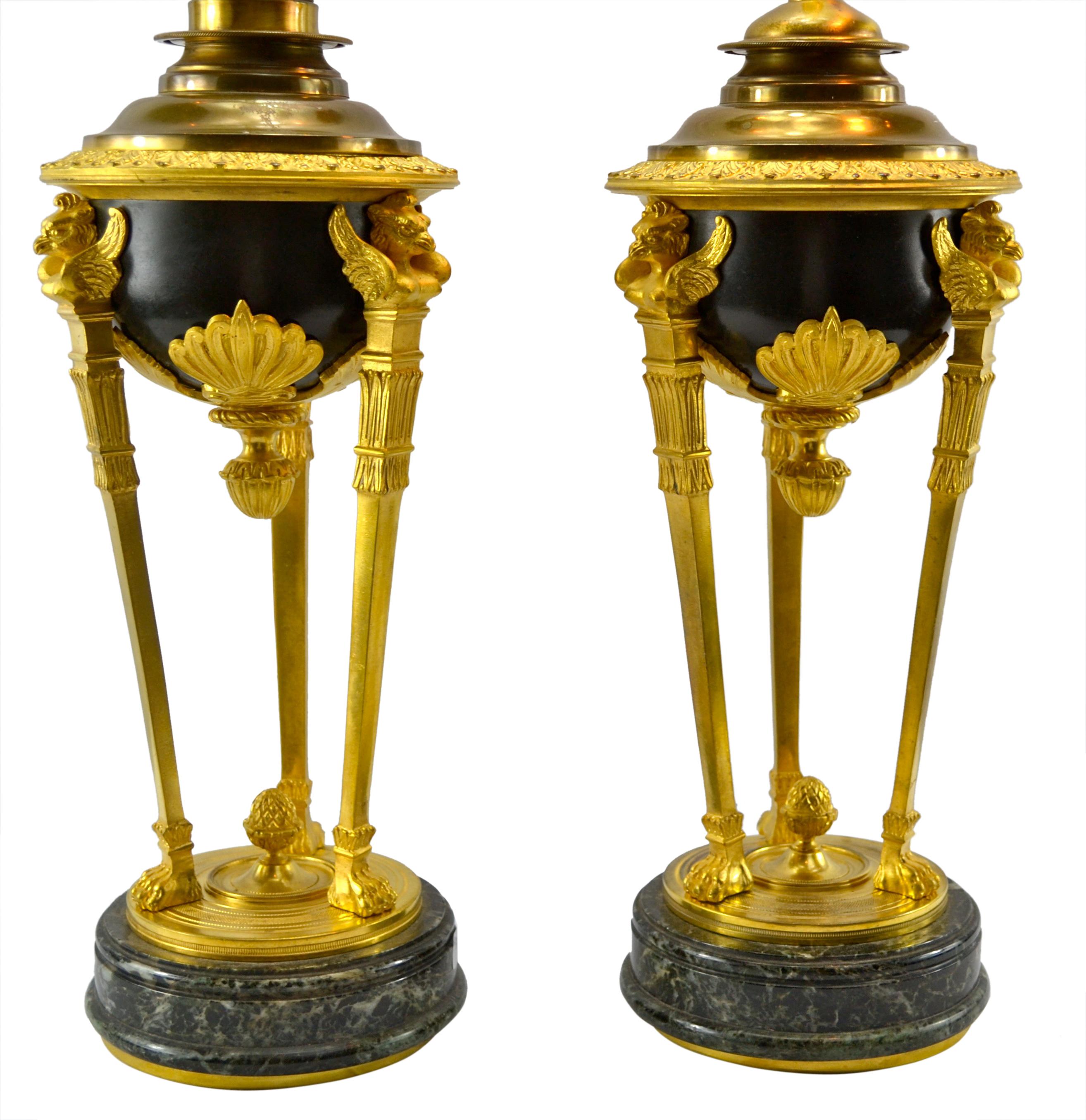 A pair of Pompeian Empire style oil lamp shaped lamps in gilded and patinated bronze on round marble bases. The lamps feature a cassolette sbody supported by three gilded columns topped with a winged eagle and ending in lion’s paw feet on a further