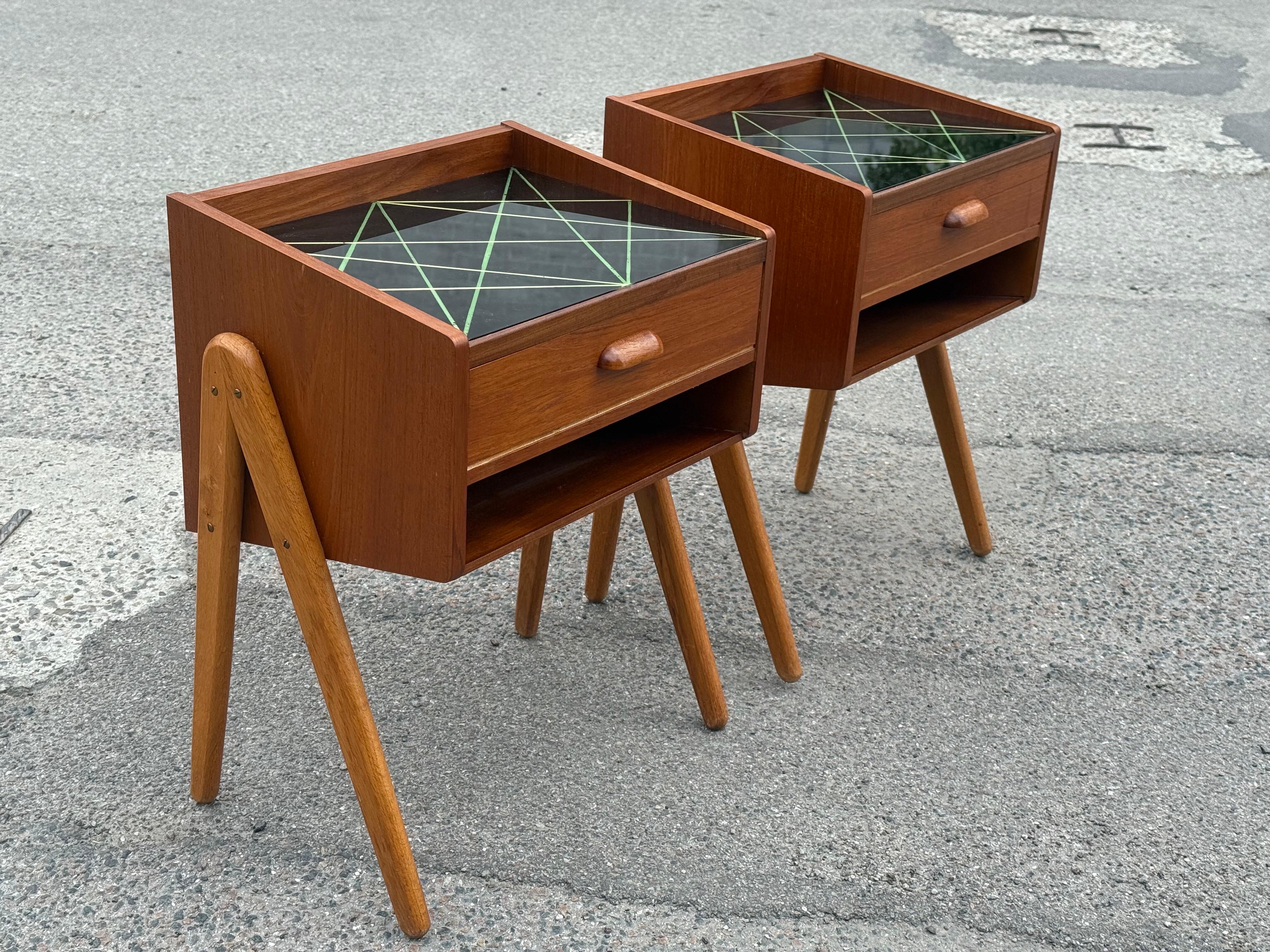 Charmante paire de tables de nuit danoises en teck des années 1960, dotées de pieds en chêne massif et d'un remarquable plateau en verre noir avec des détails géométriques verts. Chaque support comprend un seul tiroir avec une poignée en bois