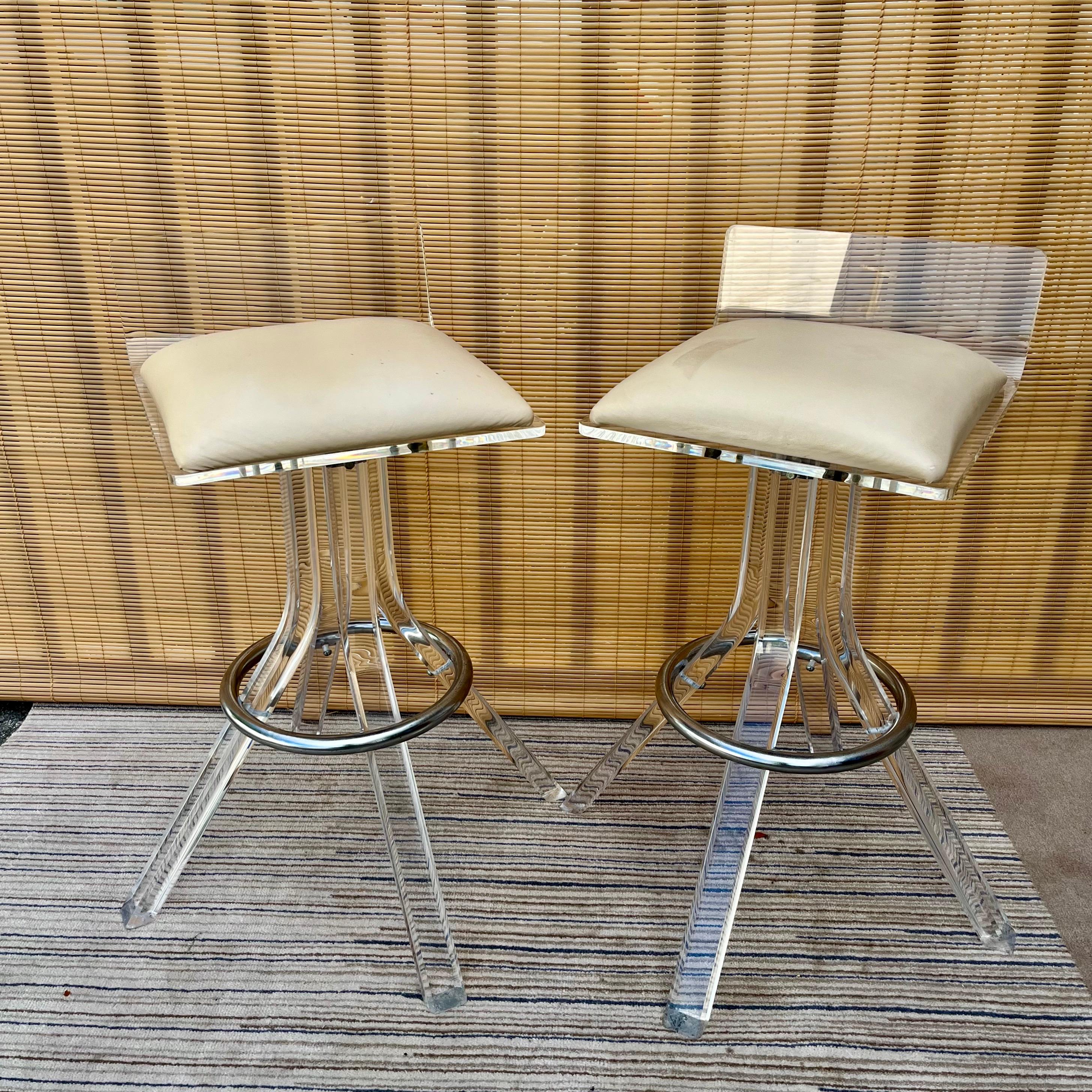 A pair of Mid-Century Modern Swivel Lucite Bar Stools in the Charles Hollis Jones' Style. Circa 1970s 
Feature swivel seats with a low bent backrest, a lucite rectangle bars pedestal with chromed steel footrests, and the original cream faux leather