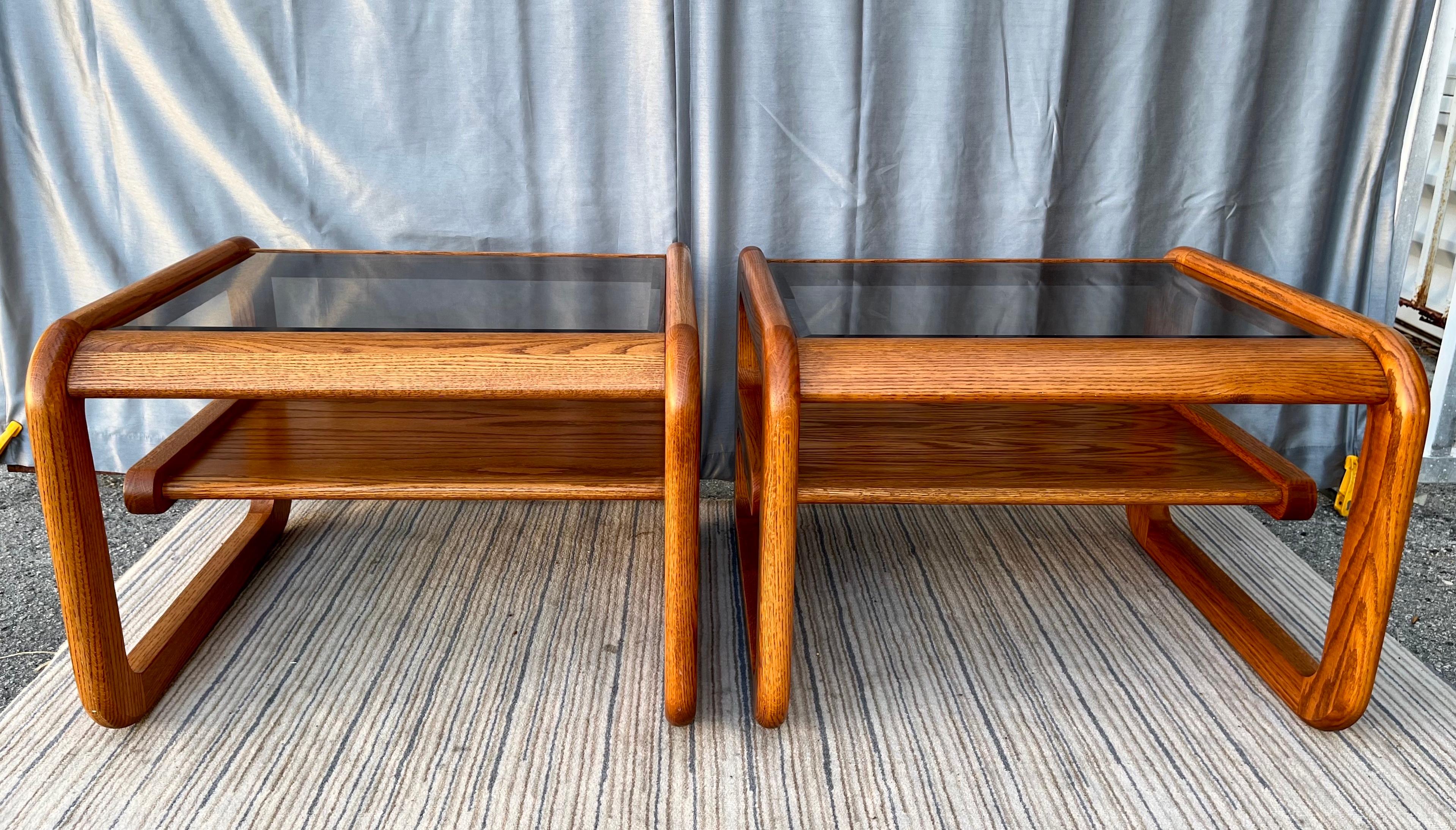 A pair of vintage mid century modern two-tier smoke glass top side tables in the Lou Hodges Style. Circa 1970s
Feature two level oak square frames with lightly smoked beveled glass insert, rounded edges, a cantilever intermediate shelf, and a